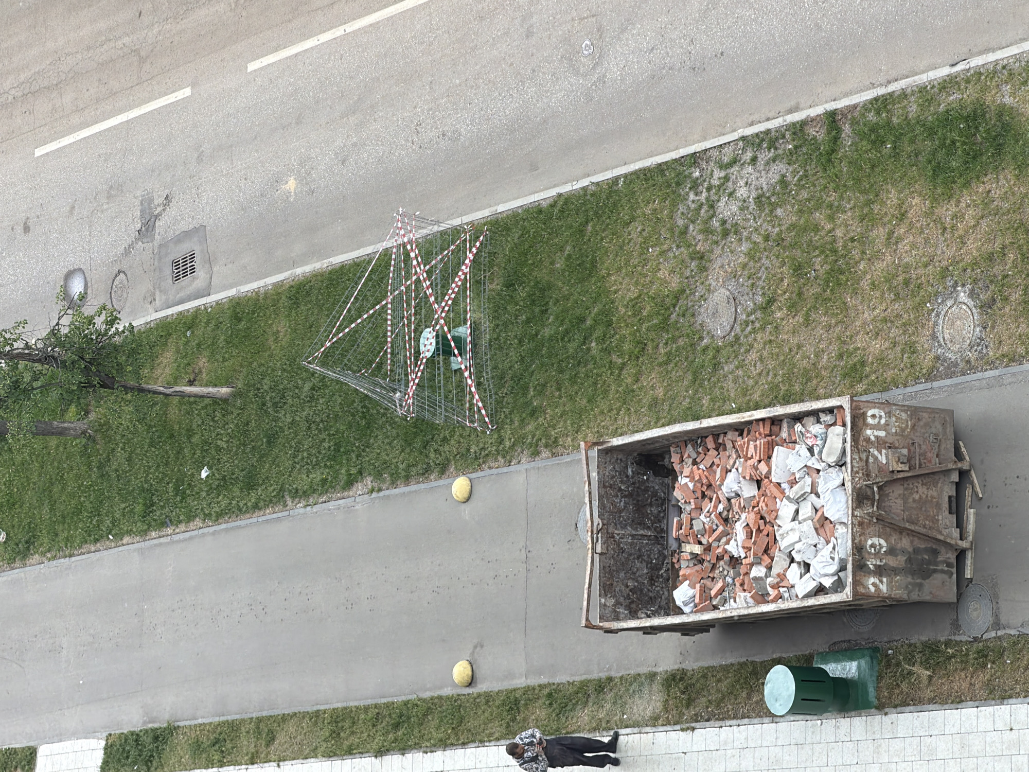 The image presents an aerial view of an urban scene. A two-lane road is visible at the top of the frame. Adjacent to the road is a grassy area containing a safety barrier composed of metal scaffolding and red and white striped tape. Alongside the grassy area is a pedestrian walkway on which a filled dumpster is situated, containing construction debris consisting of red bricks and white debris. A person wearing dark clothing is lying prone on the sidewalk beside the dumpster. A green bollard is located near the dumpster. The location is likely Moscow, Russia.