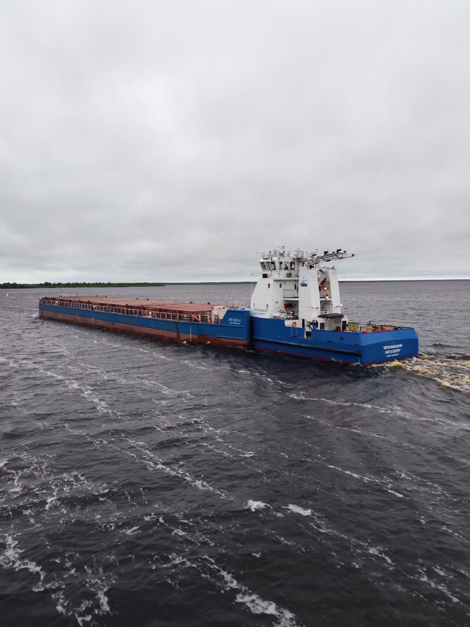 A wide, eye-level outdoor shot depicts a blue and white river tugboat pushing a barge across dark, choppy water under a gray sky. The tugboat is positioned on the right side of the barge. The barge appears to be carrying a dark-colored load. The name “ЧЕРЕПОВЕЦКИЙ МЕТАЛЛУРГ” is visible on the back of the tugboat. The scene takes place on a body of water; what appears to be land is visible in the distance on the horizon, including a single sailboat. This is likely taken in Bereg, Russia.