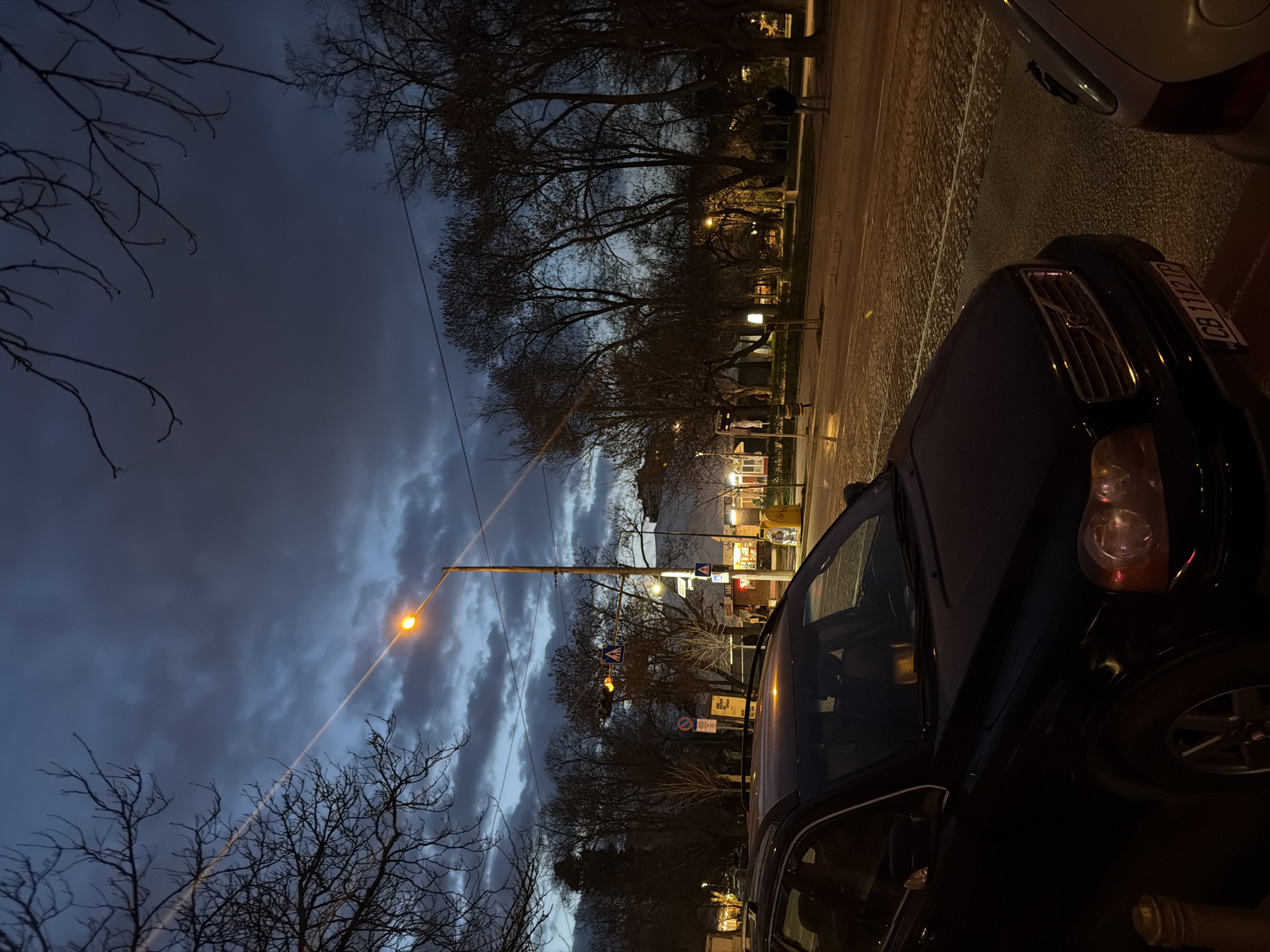 This image captures an urban street scene at dusk or early night in Sofia, Bulgaria, confirmed by the "CB" prefix on the visible license plate. The sky is heavily clouded with dark blue-grey tones, though a lighter blue is visible near the horizon. Artificial lighting dominates the scene.
In the foreground, a dark-colored (likely black) Volvo XC90 SUV is parked on the left, facing right. Its front is partially obscured by the camera's angle, but the Bulgarian license plate "CB 1112 TP" is clearly legible on the front bumper. To the immediate right of the Volvo, the rear quarter panel and taillight of a light-colored vehicle are partially visible. A brass-colored bollard is positioned in the bottom-left corner.
The road surface, stretching from the midground to the background, appears to be a mix of asphalt and cobblestones, reflecting ambient light, possibly due to dampness. A tall lamppost with an illuminated orange-yellow light stands centrally in the midground. Below it, a flashing amber pedestrian signal and a blue square sign with a white pedestrian crossing symbol are affixed to a pole. Other indistinct street signs are visible.
Bare deciduous trees, silhouetted against the sky, line the street and define a park or green area on the right side of the image. Further back, lights from commercial establishments or residential buildings illuminate the background. One individual, dressed in a dark jacket, is visible walking away from the camera on a path within the park area on the right. A yellow utility or post box is visible on the far right pavement. A faint, diagonal light streak is present in the upper left quadrant of the sky.