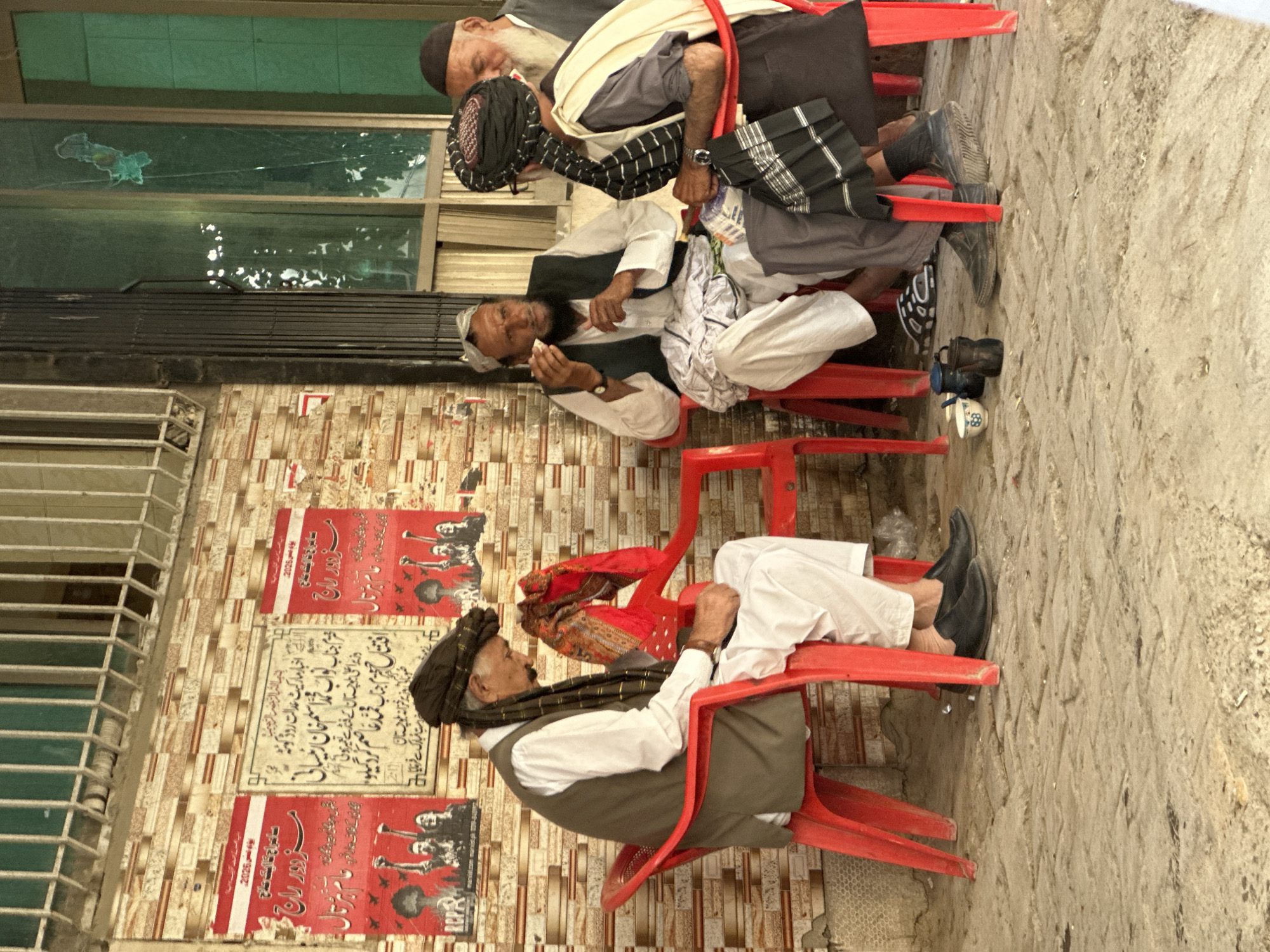The image shows three men sitting on red plastic chairs along a wall in Quetta City Tehsil, Pakistan. The wall is adorned with patterned tiles and two red posters featuring images and text in Urdu, with the text "Muzdoor Raj" visible. The men are dressed in traditional clothing; the man closest to the camera wears a black turban with a patterned scarf, a white shirt, and a vest, while the others are in turbans and similar attire. One of the men appears to be eating, while the others are seated. The background includes a window and a narrow street or walkway.