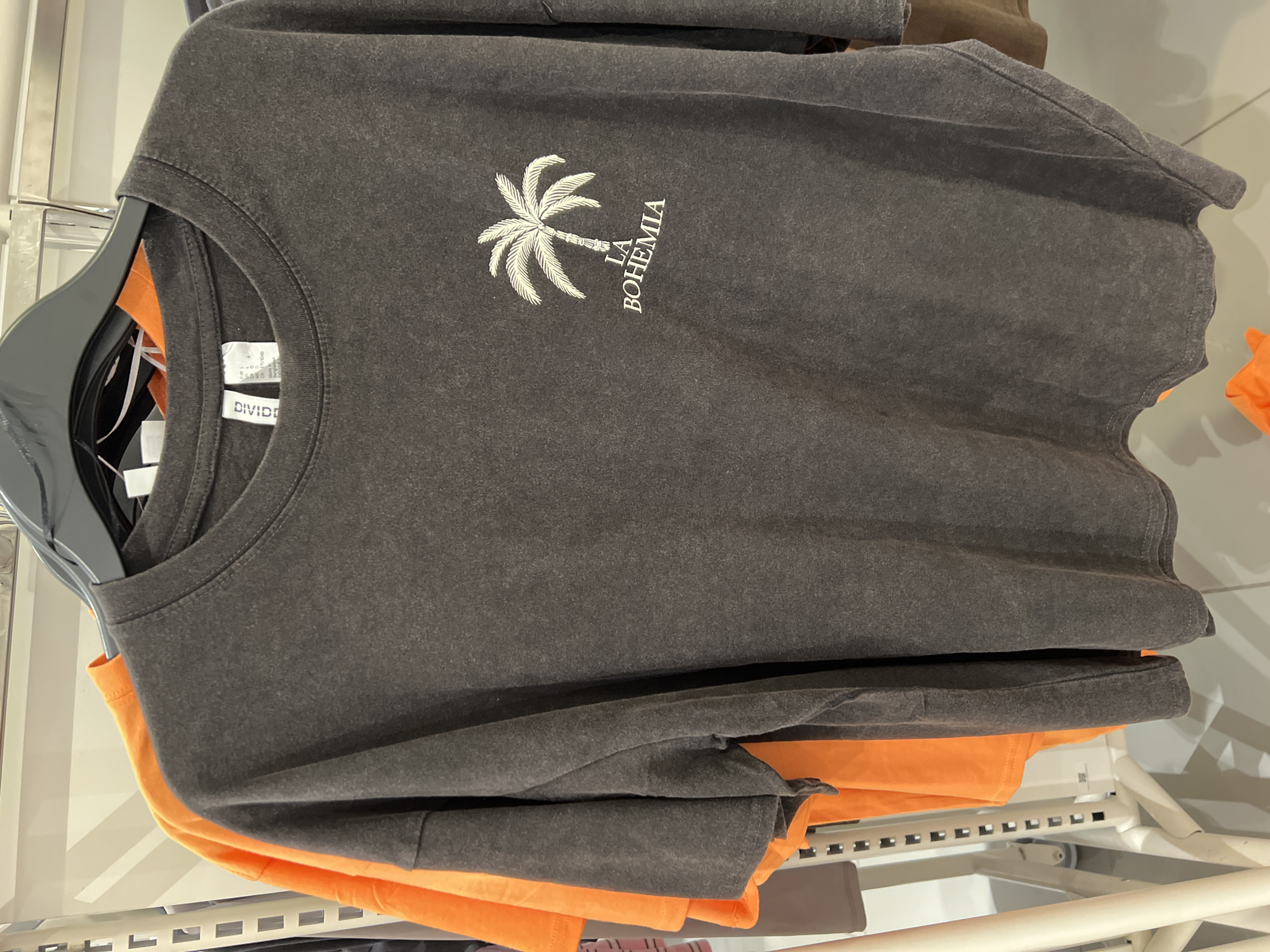 The image shows a dark gray t-shirt with a white graphic of a palm tree and the text "LA BOHEMIA" printed on it. The t-shirt is hanging on a black hanger, with a bright orange t-shirt visible behind it. The background suggests a retail store environment, with clothing racks and lighting. The t-shirt appears to be made of a washed or distressed fabric. No people or specific landmarks are visible. The image does not provide any information about the location in the city of Auburn, United States of America.