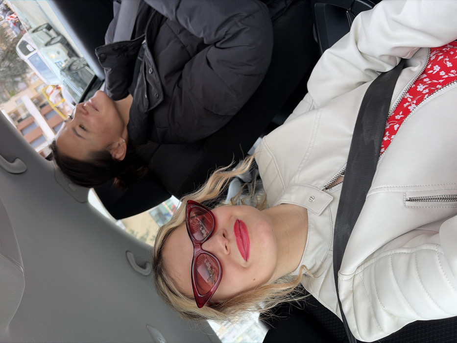 The image depicts the interior of a vehicle traveling on a street in Budva, Montenegro. In the foreground, a young female, with blonde, wavy hair, is seated wearing red cat-eye sunglasses and bright red lipstick. She is dressed in a light-colored, possibly white or off-white, quilted leather-style jacket over a red floral patterned garment, with a dark seatbelt secured across her chest. Her head is turned slightly towards the left, and she is looking directly at the camera with a slight smile. In the mid-ground, to her right, another young female with dark hair pulled back is seated, wearing a black puffer jacket. Her face is illuminated by sunlight, and she is looking towards the right, appearing relaxed with her eyes closed or squinted. Through the vehicle's windows, an urban street scene is visible, featuring multi-story buildings with balconies, other vehicles including a white SUV and a dark sedan, and bare trees under bright daylight conditions.