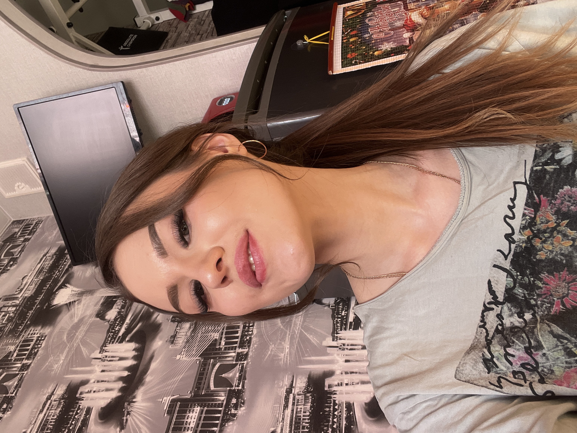 A woman is lying on a patterned surface resembling a city skyline, wearing a light grey t-shirt with a dark floral and script print. She has long, brown hair and is wearing hoop earrings and a delicate necklace. Her face is tilted towards the camera, displaying full makeup with extended eyelashes and pink lipstick. In the background, there's a computer monitor, an exercise machine, and a calendar on a black surface. It is not possible to identify a specific location in Noviy, Russia from this image.