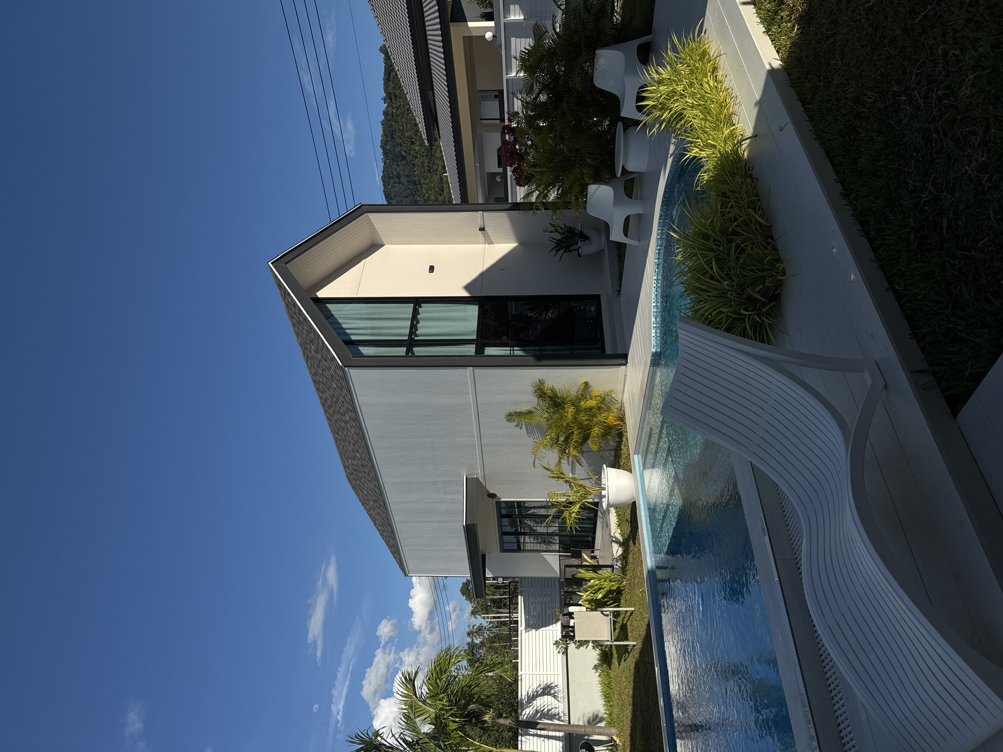 This is a photograph taken in Baan Tai, Thailand, featuring a modern house with a swimming pool. The image is oriented vertically.
The main subjects are the house and its surroundings. The house is a contemporary structure with a light-colored facade, dark-framed windows, and a dark roof. There is a swimming pool adjacent to the house, filled with clear blue water. White outdoor chairs are placed near the pool. Green vegetation, including palm trees and other lush plants, surrounds the pool and house, indicating a tropical setting. In the background, a lush green hillside is visible, suggesting the location is near mountains. Overhead, a bright blue sky with a few wispy clouds is present, and power lines are visible in the upper left portion of the image.
The photograph appears to be taken during the daytime, under clear and sunny weather conditions, as indicated by the bright sunlight and blue sky. The overall scene conveys a sense of relaxation and luxury, typical of a tropical resort or private villa. No people are visible in the image, and no text is present. The arrangement of the house, pool, and landscaping suggests a well-maintained property designed for outdoor living.