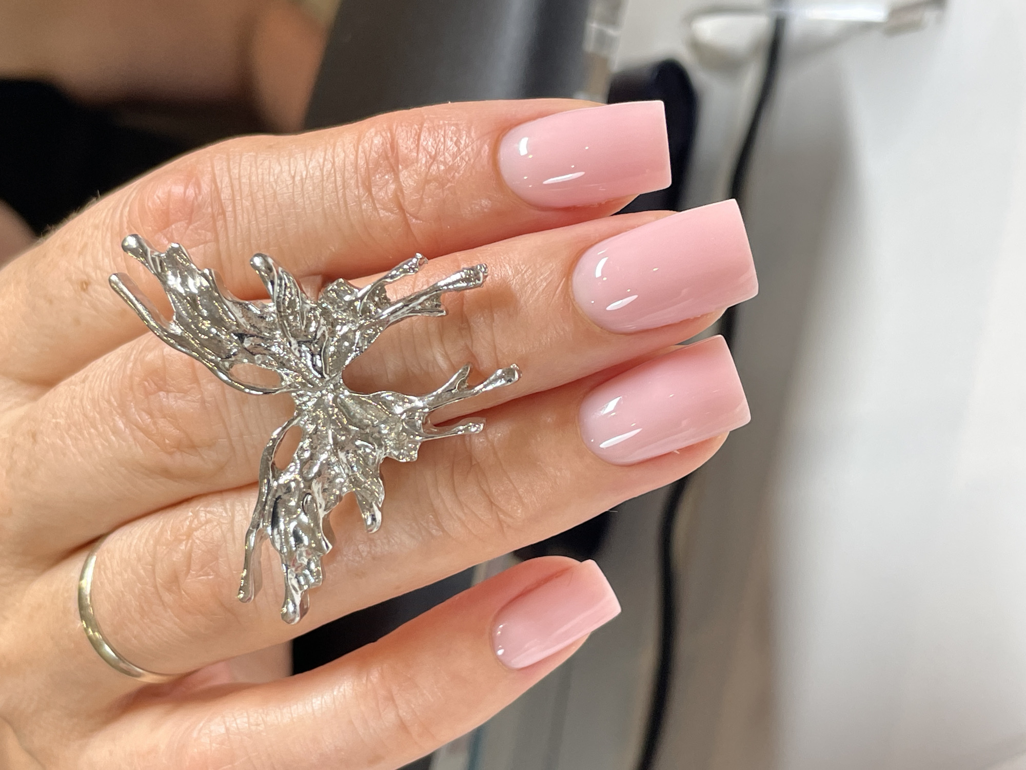 A close-up photograph captures a hand adorned with manicured nails and jewelry. The nails are square-shaped and painted a glossy, light pink. A large, silver ring in the shape of an abstract butterfly is worn on the index finger. A simple gold band is on the pinky finger. The hand is resting against a dark gray surface, with a blurry white object in the background. The environment appears to be indoors, possibly a salon or studio. There is no evidence to suggest this image was taken in Izhevsk, Russia.