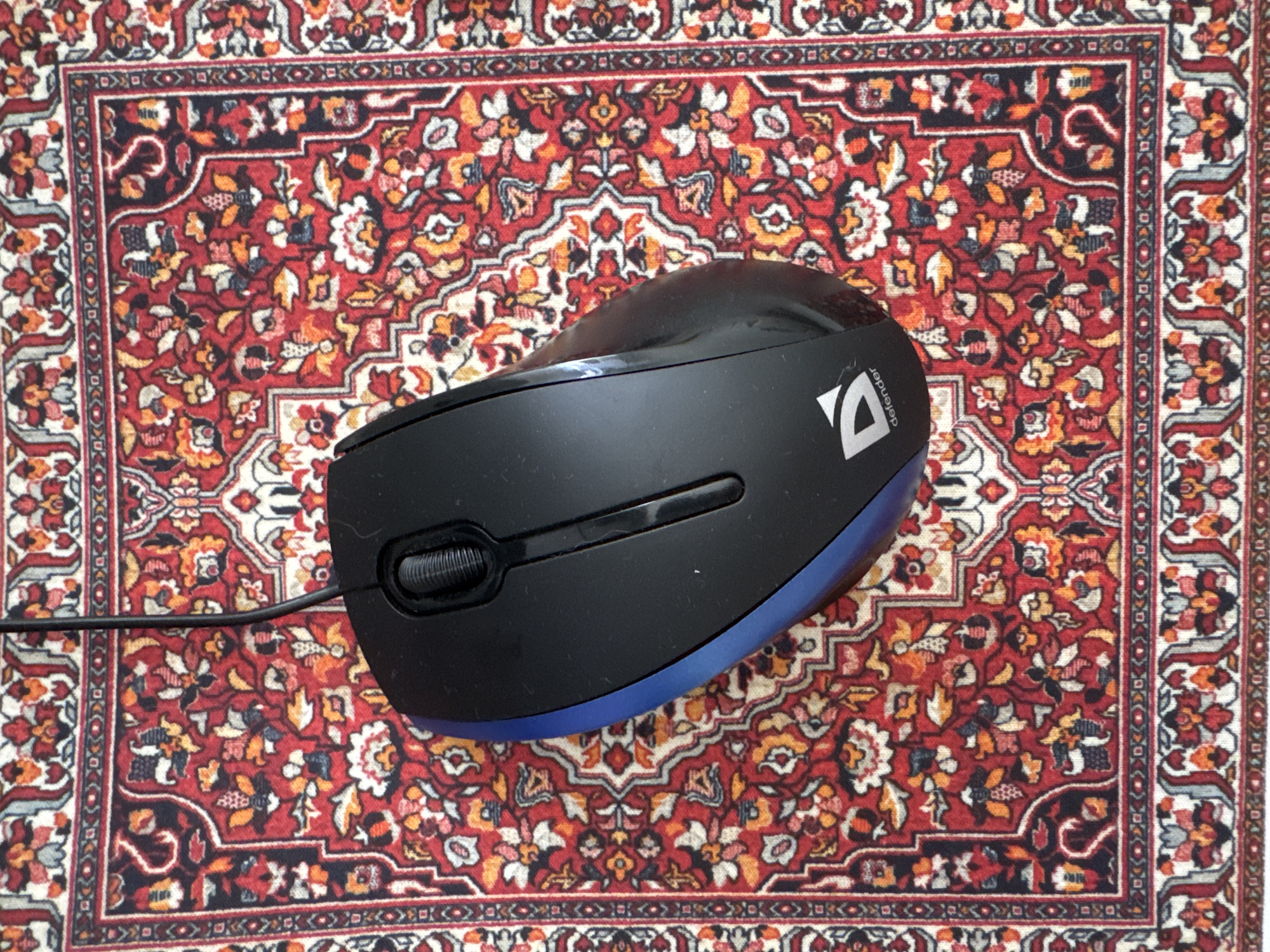 A black computer mouse with a blue trim is positioned on a red oriental rug with intricate patterns. A black wire extends from the top of the mouse. The rug is adorned with floral motifs in shades of red, white, blue, and orange. The mouse has a scroll wheel and a logo that reads "defender". The image is taken from a top-down perspective. The scene is indoors, on a flat surface. There are no people or animals visible in the image. The specific location in Боголюбово, Russia is not discernible from the provided image.