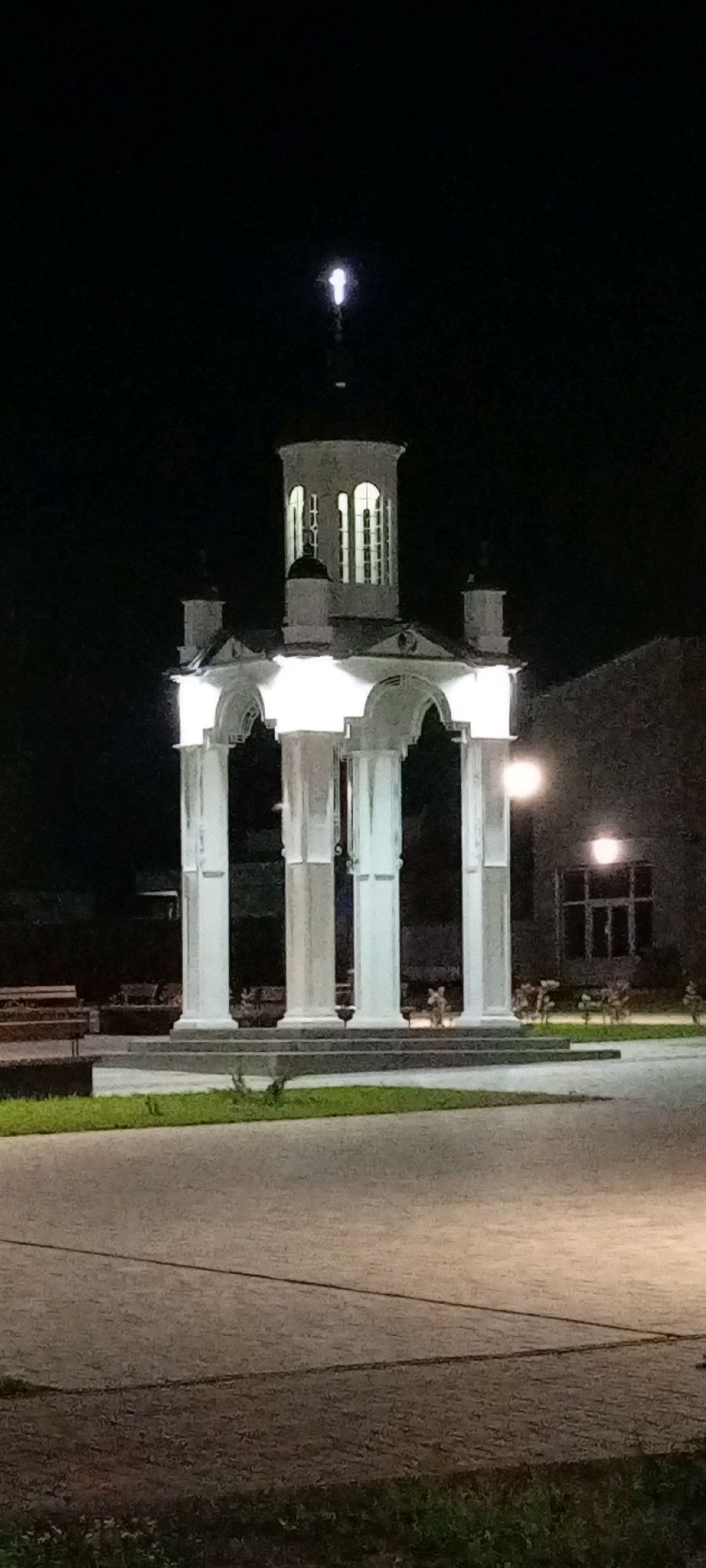 The image is a nighttime photograph of a white, illuminated monument located in the city of Kirsanov, Russia. The monument features a circular base with four large, white columns supporting an arched structure. Above this, there is a multi-tiered, octagonal tower with arched windows, topped with a smaller dome or spire. The monument is lit from below by bright white lights, making it stand out against the dark background. A bright light source, possibly a streetlamp, is visible on the right side of the image, illuminating a building and some plants. The foreground shows a paved area and some grass. The scene appears to be a public space, possibly a park or square, given the presence of benches visible on the left side of the monument. There are no people visible in the image. The overall impression is one of quiet illumination and architectural presence in the dark.