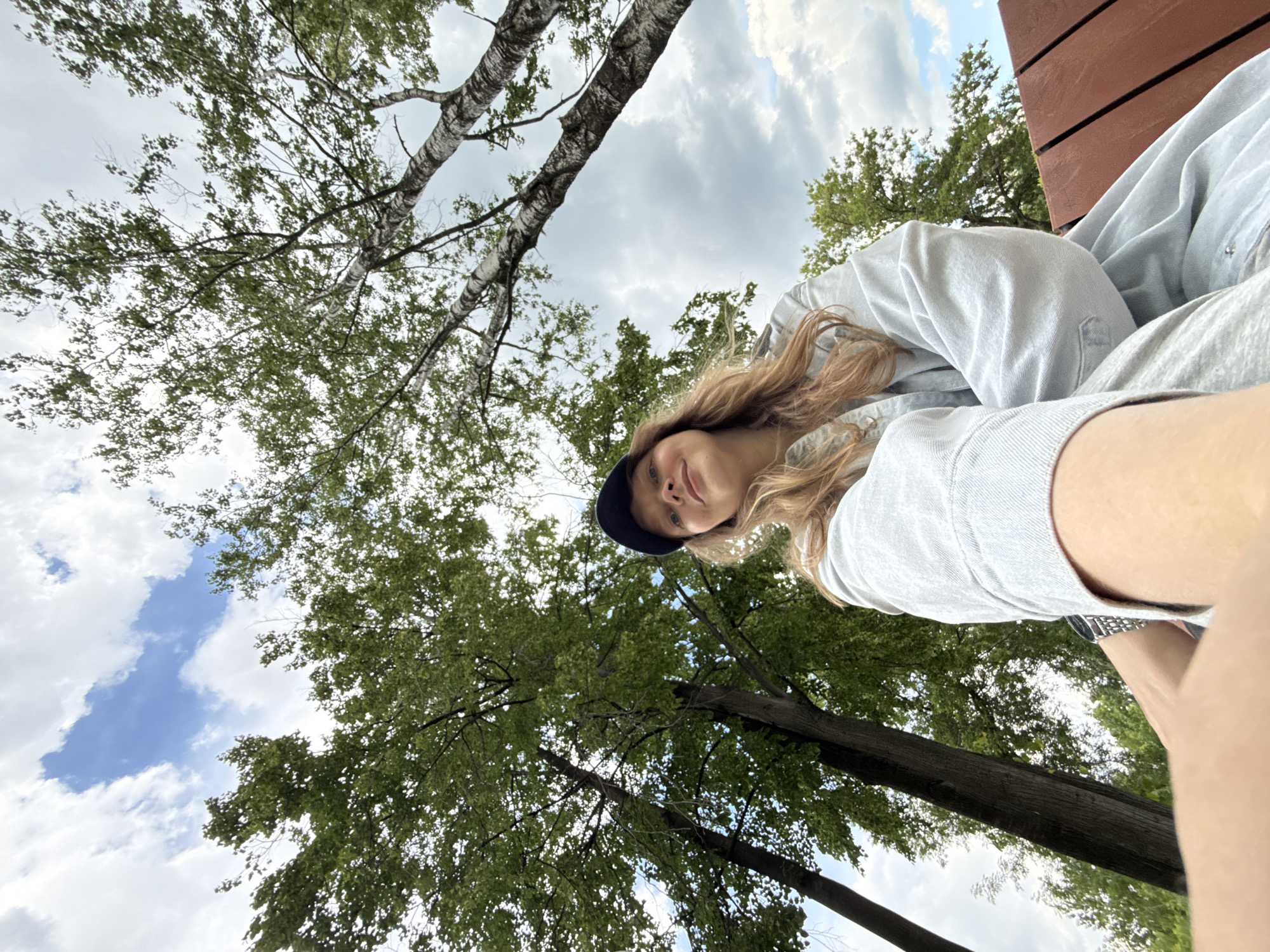 A young woman wearing a light blue denim jacket and a black baseball cap takes a selfie outdoors. She has long, blonde hair and is smiling slightly. She is sitting on a reddish-brown wooden bench, and behind her, tall trees with green leaves reach towards a cloudy sky. The image is taken from a low angle, looking up at the woman and the trees. There are no other people or objects of significance in the frame. The location is not specified to be in Izhevsk, Russia.