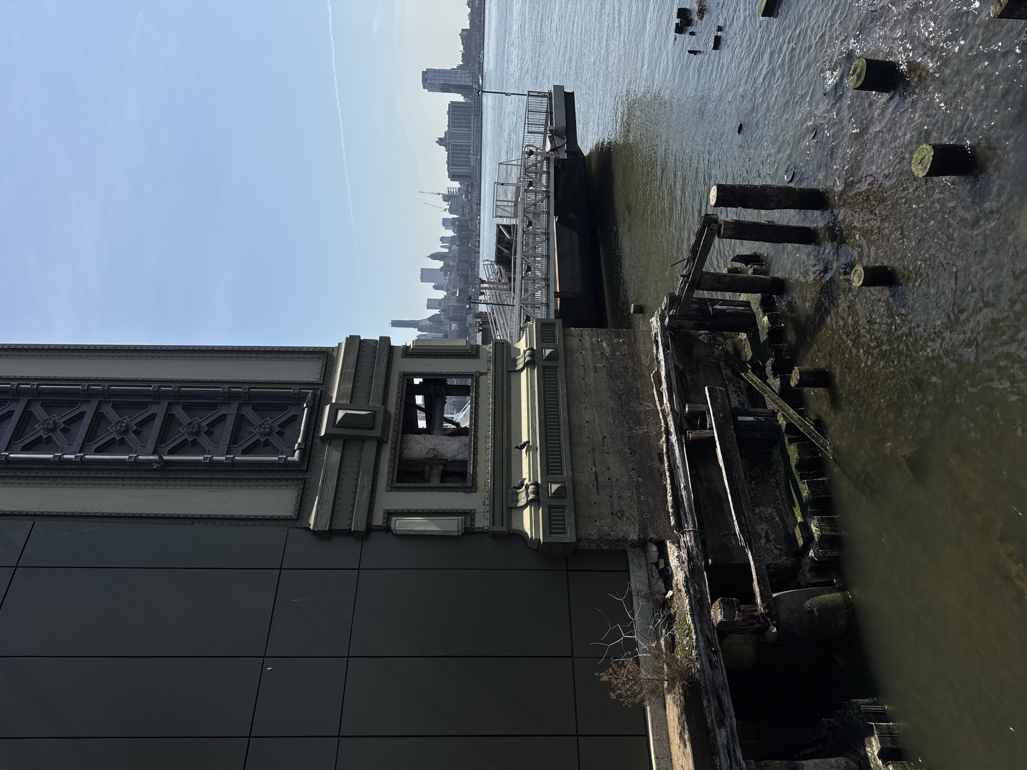 The image depicts a waterfront scene in Brooklyn, New York City, United States of America, specifically near the Manhattan Bridge, looking west across the East River towards the Manhattan skyline.
The left side of the frame is dominated by two structures: a modern building faced with dark grey, rectangular panels, and an adjacent, ornate bridge tower. The tower, part of the Manhattan Bridge, is dark green-grey, constructed from riveted metal plates with decorative cross-bracing and a concrete base. Two dark birds are perched on a lower ledge of this tower. Below the modern building and the tower, a small patch of dry earth supports a dry, shrub-like plant with bare branches.
In the foreground, numerous dilapidated wooden pier remnants are visible, including large horizontal beams and vertical pilings protruding from the dark, rippling water of the East River. Some pilings are partially submerged and covered with green moss or algae.
In the mid-ground, a modern grey metal ferry dock structure, complete with gangways and railings, floats on the water, connected to the land near the bridge tower. A small lamppost stands on the dock.
The background features the distinctive silhouette of the Manhattan skyline under a clear, light blue sky. Numerous skyscrapers and residential buildings are visible, some with active construction cranes. A faint white contrail from an aircraft stretches horizontally across the distant sky. No people are present in the scene.