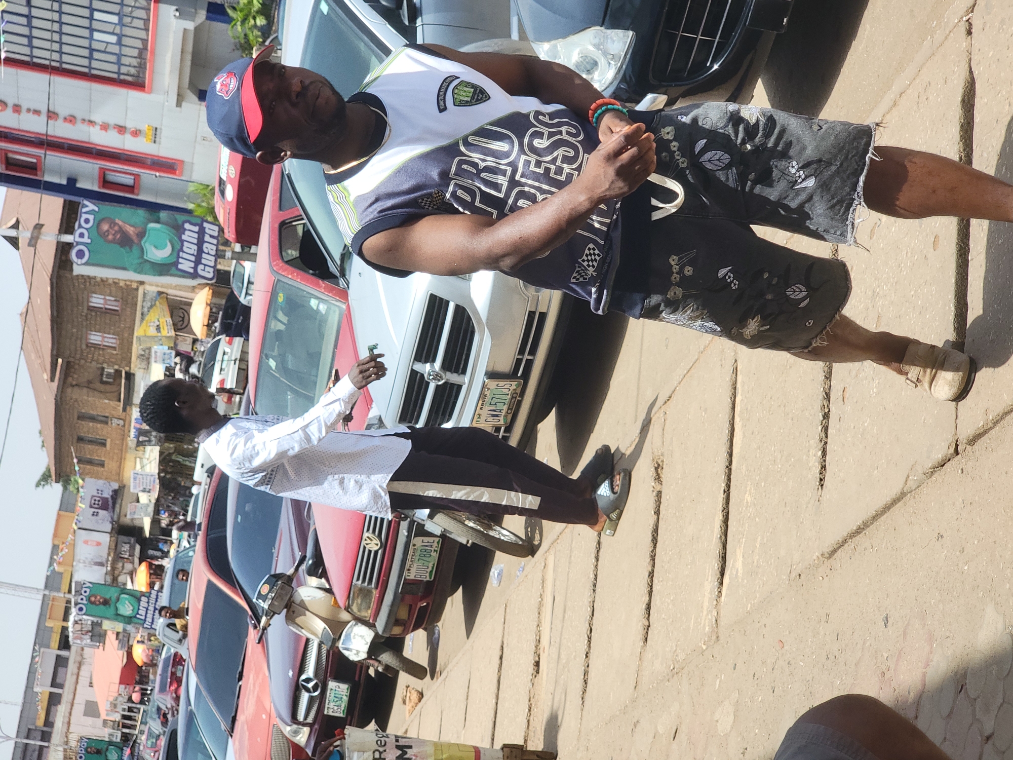 The image shows a street scene in Jos, Nigeria, characterized by several vehicles and pedestrians. In the foreground, two men stand beside a light-colored car. One man is wearing a baseball cap, a white and blue t-shirt that reads "PRO-RESS," and printed shorts. He is positioned in front of the car, facing right with his hands near his chest. The other man, wearing a white shirt and dark trousers, is to his left and slightly behind him, gesturing towards the front of the vehicles.
A row of cars, including a red one, lines the street. In the background, commercial signage is visible on nearby buildings, including an "Opay" advertisement. A small motorcycle is parked near the red car. A glimpse of other pedestrians and storefronts suggests a busy commercial area. License plates from Plateau and Abuja are visible on the cars, confirming the location within Nigeria.