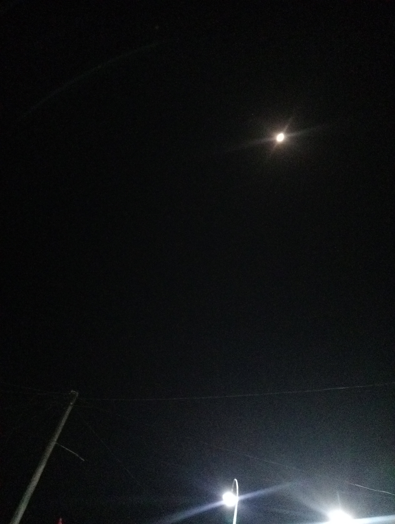 The image captures a night sky with a crescent moon visible in the upper right quadrant. The sky is predominantly dark, with some grainy texture. In the lower portion of the image, two streetlights are brightly illuminated, casting beams of light upwards and outwards. A power pole or utility pole is visible on the left side of the frame, with thin wires extending horizontally across the sky. The scene appears to be an outdoor urban or semi-urban environment at night. There are no people or discernible actions depicted in the image. The location is identified as Rasuan Baru, Indonesia.
