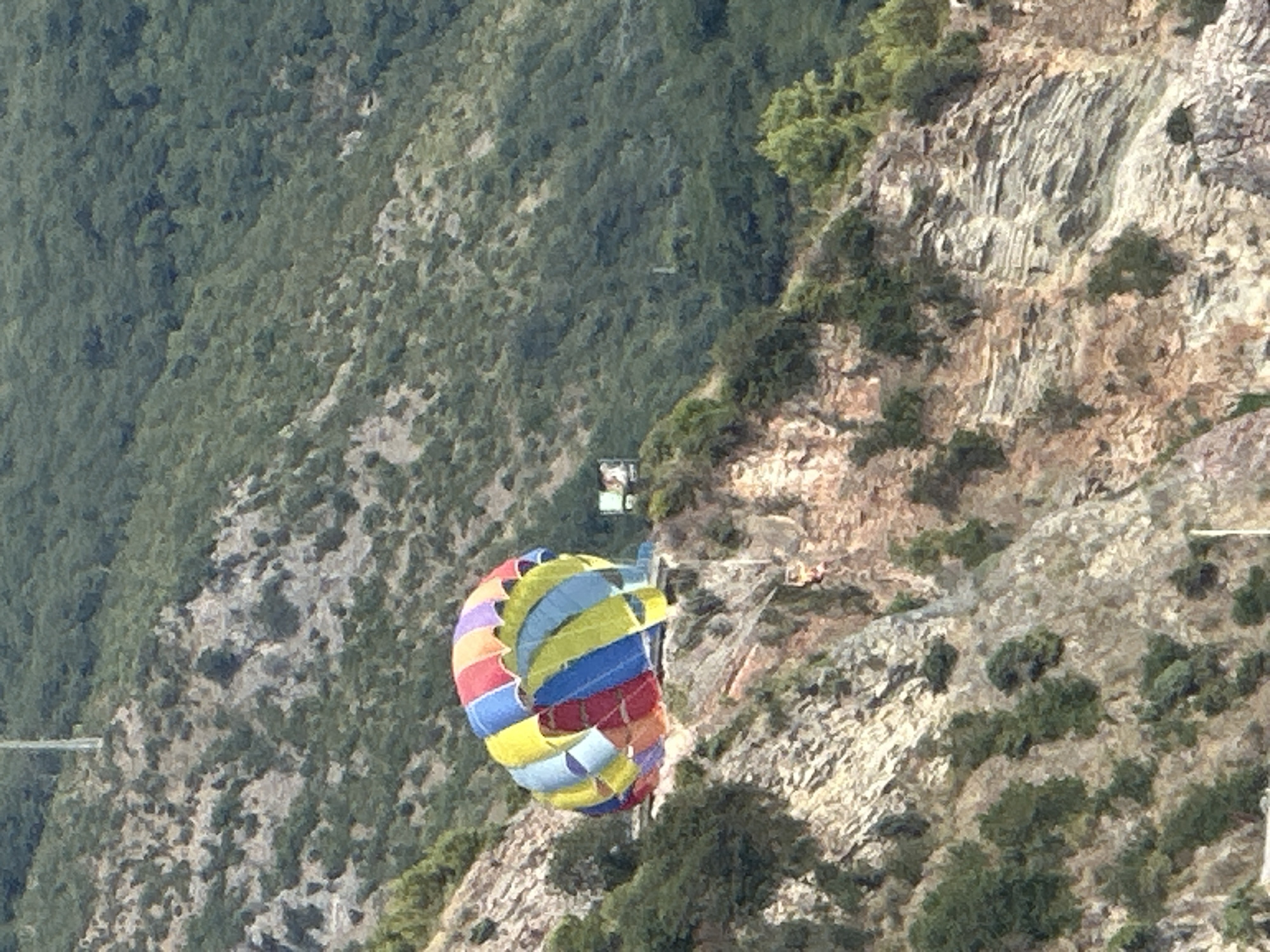 In Rafailovići, Montenegro, a multi-colored parachute, likely for parasailing, rests on a cliff edge overlooking a densely vegetated mountainside. The parachute, composed of vibrant panels of yellow, blue, red, and purple, appears deflated or partially deployed, its fabric billowing outwards. The cliff face is rugged and rocky, with sparse green vegetation clinging to its slopes. In the background, the mountains are covered in thick green foliage. A small, distant structure, possibly a building or a sign, is visible on the cliff face to the right of the parachute. The scene suggests a tourist activity site, possibly a launch or landing area for recreational flights.