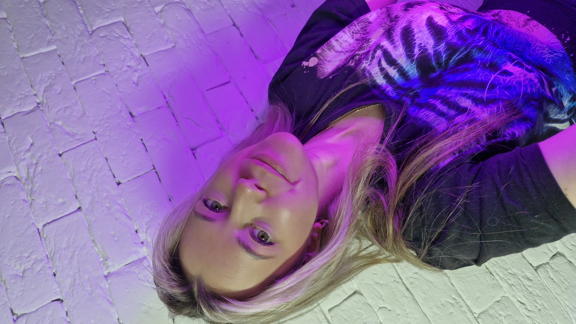 A young woman is depicted lying on a white, brick-patterned surface, looking directly at the camera. Her head is positioned in the lower-center of the frame, and her body extends diagonally upwards and to the right. She has long, blonde hair that spreads around her head on the surface, and bright green eyes. A faint, calm smile graces her lips.
She is wearing a dark, possibly black, t-shirt with a vibrant and intricate design in electric blue and purple, resembling glowing tiger stripes or a tie-dye pattern, which is prominently lit by the colored lights. A thin gold chain necklace is visible around her neck. The entire scene is bathed in strong artificial lighting, predominantly purple, which casts a vivid magenta hue over the white bricks and illuminates sections of her hair and face. Patches of blue light are also evident, particularly on her t-shirt.
The setting appears to be an indoor space, such as a studio or a room with decorative lighting, given the intense and colored illumination. The activity is a posed portrait. No time of day or specific weather conditions are discernible due to the artificial lighting. There are no explicit visual cues in the image itself to indicate the location as Otchor, Russia. No text is visible.
