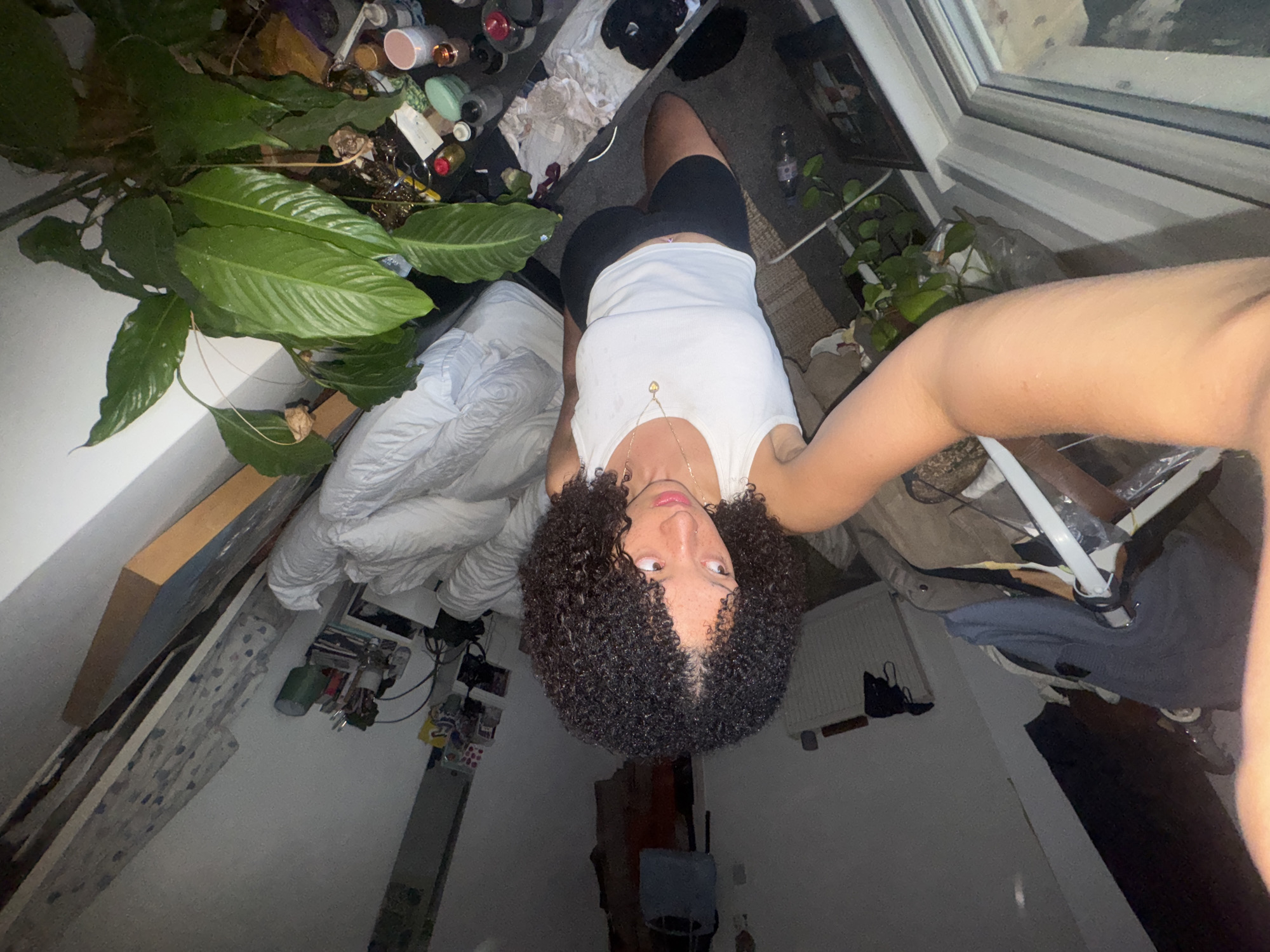 A high-angle, wide-lens self-portrait captures a person looking up towards the camera in what appears to be a bedroom or living space. The subject, positioned centrally, has dark, curly hair and is wearing a white ribbed tank top and black shorts. A delicate gold necklace with a small pendant is visible around their neck, and they have a nose piercing. Their gaze is directed upwards at the camera, held by an arm partially visible on the right.
The room is characterized by its cluttered, lived-in appearance. To the left of the subject, a large potted plant with broad green leaves dominates the foreground, beside a crumpled white duvet or comforter. Further left, several art pieces or canvases are leaning against a light-colored wall. Above and behind the subject, a surface is densely packed with numerous small items, including bottles, jars, and other miscellaneous objects, suggesting a vanity or desk area.
To the right, a white-framed window allows for a glimpse of a blurry outdoor scene. Below the window, a white radiator is affixed to the wall, and various items of clothing are draped over what might be a chair or clothes rack. Another small potted plant is visible near the window. The floor appears to be dark carpeting or flooring, with additional scattered items. The lighting is dim, suggesting indoor artificial light or evening conditions. No specific text is visible within the image.