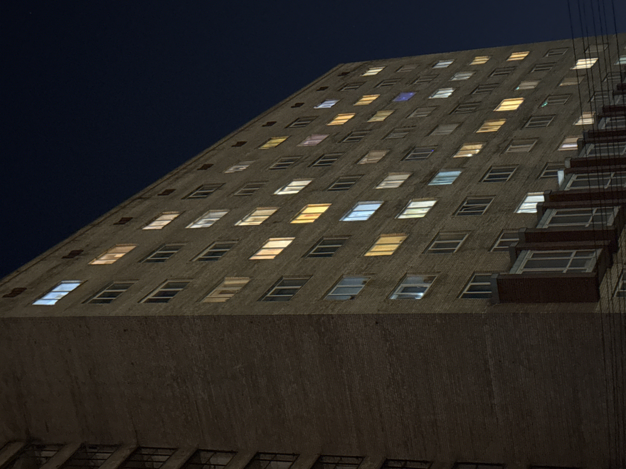 An evening low-angle shot captures a massive brutalist-style building against a deep blue, clear night sky. The imposing structure, likely constructed from concrete or dark brick, stretches upwards and diagonally across the frame from bottom left to top right. Its facade is punctuated by a grid of numerous rectangular windows.
Many of these windows are illuminated, casting a variety of light colors onto the dark exterior. Dominant light colors include warm yellows and oranges, bright whites, and cooler light blues, with a few hints of pink or purple. Some windows remain dark, creating an irregular pattern of light and shadow across the building's surface. The varied lighting suggests different activities or individual preferences within the building at night.
On the far right side of the building, a series of dark, angular structures resembling balconies or external staircases are visible, receding into the distance. The overall impression is one of a large, active urban building after dark in Saint Petersburg, Russia. The time of day is clearly night, with no discernible weather other than a clear sky. There is no visible text in the image.