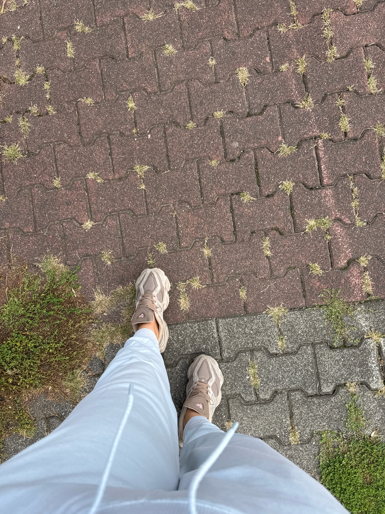 This is a first-person perspective shot of a person standing on a cobblestone path. The path is composed of interlocking, dark reddish-brown pavers with some patches of green grass growing in the cracks. To the right of the path, the pavers transition to a lighter gray color. The person is wearing light blue sweatpants and light brown athletic shoes. The scene appears to be outdoors, possibly in a park or a residential area. The image does not provide specific visual cues to pinpoint the location to Katowice, Poland.