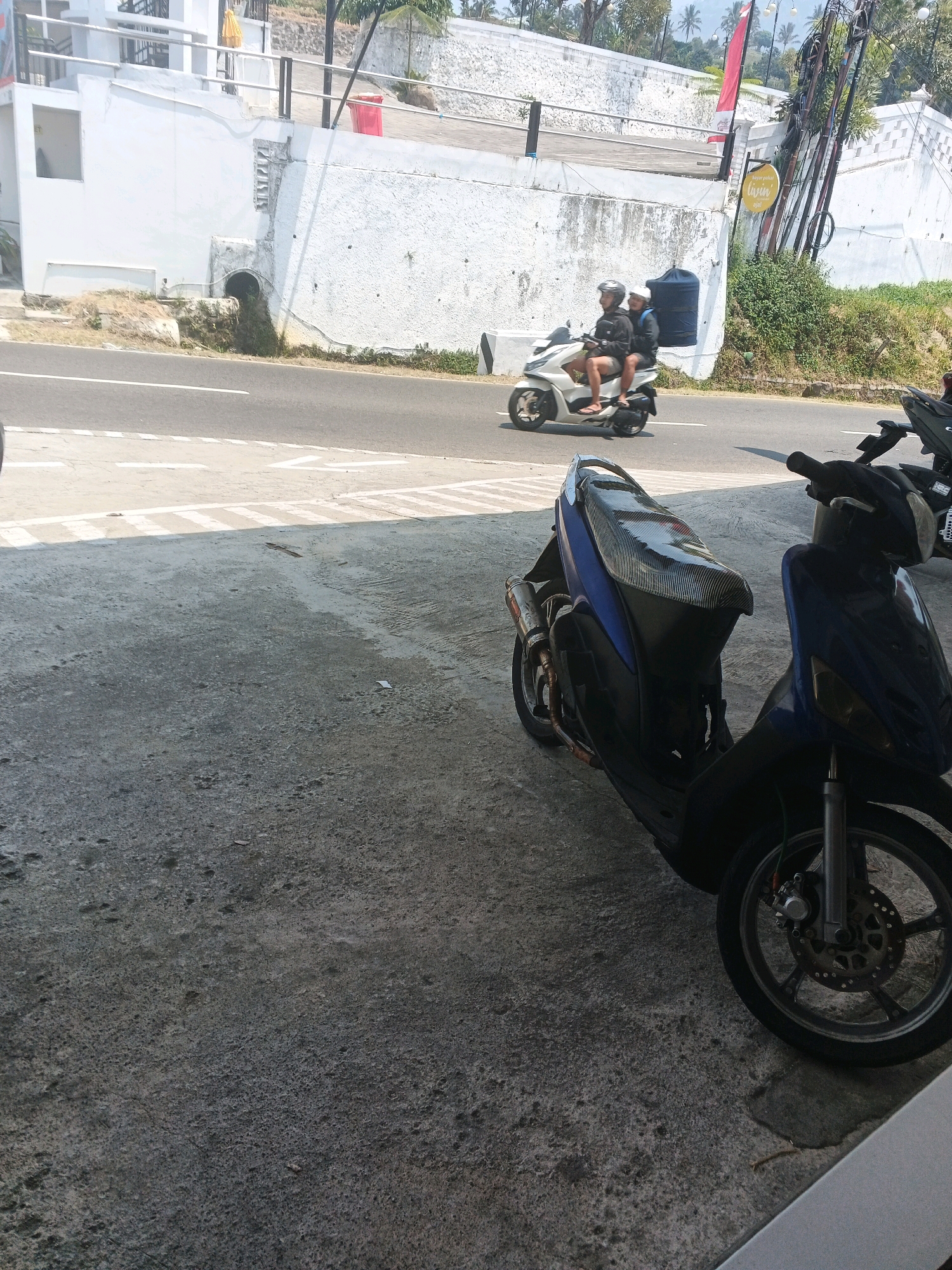 A daytime, eye-level outdoor shot shows two people on a scooter moving past a white wall. The scooter is white with two people wearing helmets. The person in the front is wearing a dark jacket and shorts, and the person in the back is wearing a dark jacket and black pants. A large black water tank is attached to the back of the scooter. The scooter is on a paved road. To the left of the road is a white wall with a drainage pipe. Behind the wall are some palm trees. To the right of the road is a green area with some trees. In the foreground, two motorcycles are parked on a concrete surface. One motorcycle is blue and the other is black. The blue motorcycle is in focus, and its front wheel, handlebars, and seat are visible. The black motorcycle is partially visible in the background. The scene appears to be located in Pakopen, Indonesia.