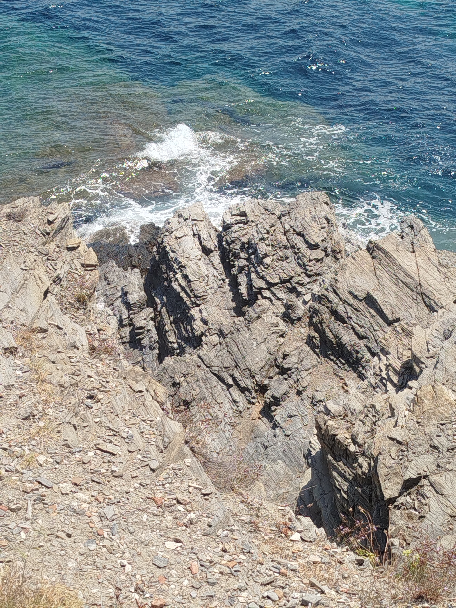 The image shows a rocky coastline with the sea in the background. The rocks are gray and jagged, with some vegetation growing on them. The sea is blue and clear, with waves crashing against the shore. The image is taken from a high angle, looking down at the coastline. The location is identified as Sithonia Municipal Unit, Greece.