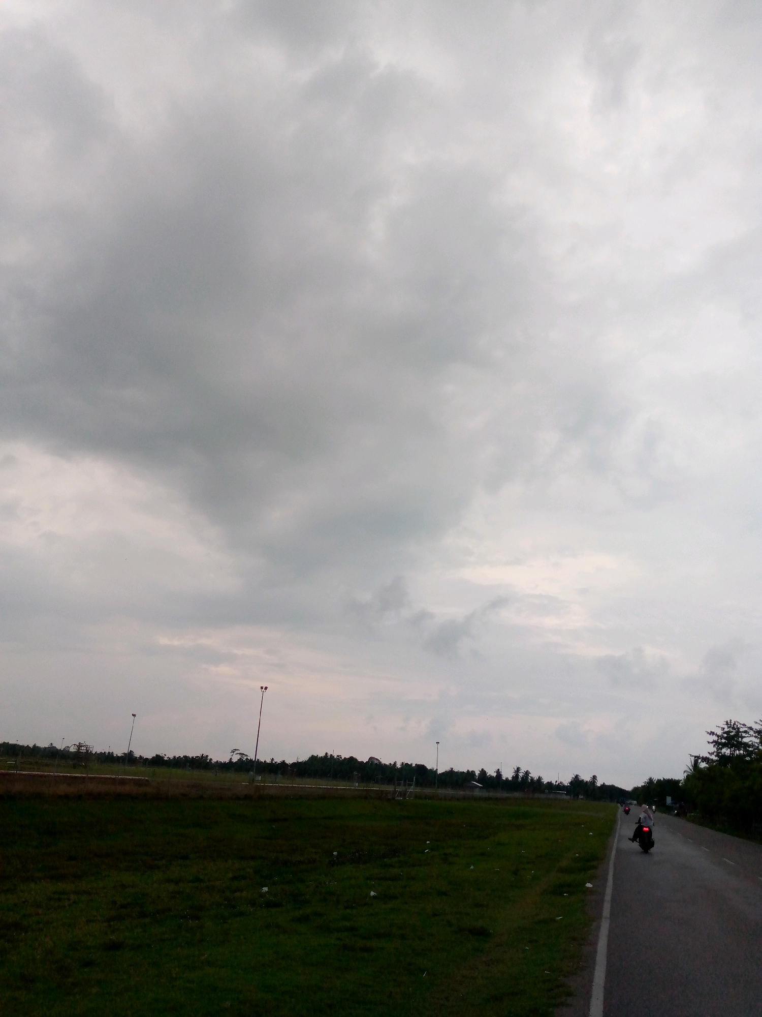 The image depicts an outdoor scene in Glokaron, Indonesia, under a cloudy sky. A paved road runs diagonally from the lower right to the middle of the image, bordered by a grassy field on the left and vegetation on the right. Several individuals are seen riding motorcycles on the road, heading away from the viewer. The field appears to be a sports field or an undeveloped area, with tall lampposts visible in the distance. The overall atmosphere is subdued due to the overcast weather.