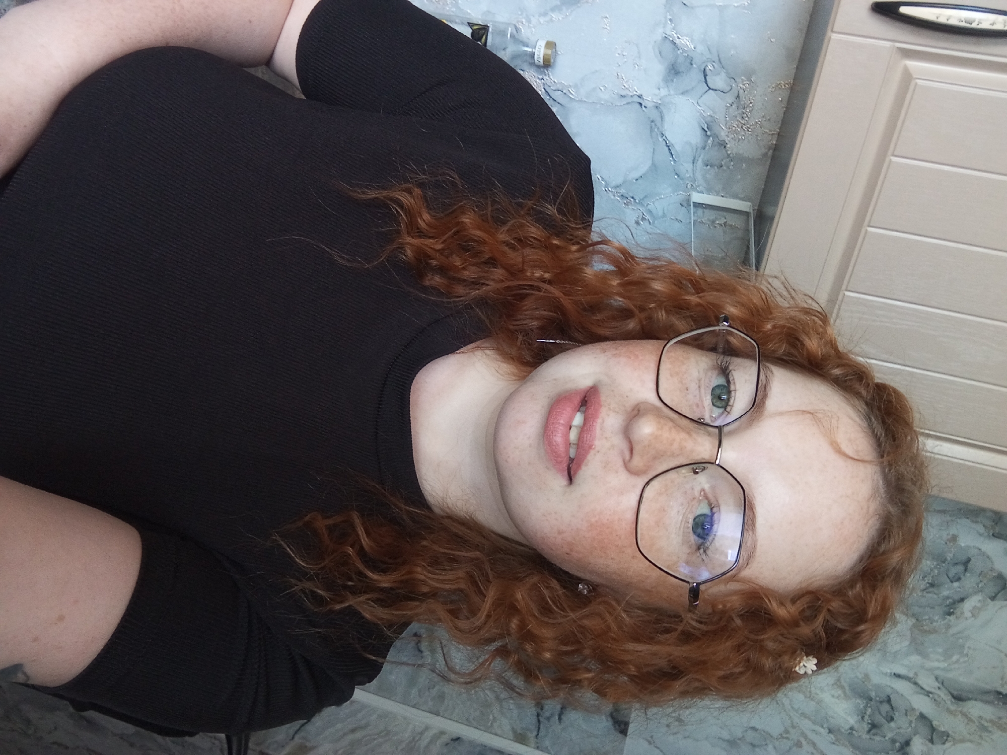 The image captures a young woman lying down and looking directly into the camera. She has fair skin with a distinctive pattern of numerous freckles across her face. Her long, curly, reddish-brown hair is spread around her head. She wears large, hexagonal-shaped eyeglasses with thin metal frames, through which her light-colored eyes are visible. Her lips are slightly parted, revealing some teeth, and are colored with a natural or light pink shade. A small white flower accessory is tucked into her hair near her right ear. She is dressed in a black, ribbed, short-sleeved top or dress.
The setting appears to be indoors, with her head resting on a surface featuring a light blue and grey marble-like pattern. In the upper right background, a light-colored wooden cabinet or furniture piece with horizontal paneling is visible. Some indistinct markings or text can be seen on the cabinet handle. Towards the upper left, partially obscured by her hair and shoulder, is a small glass bottle with a cork stopper. The label on this bottle distinctly shows the Cyrillic text "РУССКАЯ ВОДКА" (RUSSIAN VODKA), providing a clear location cue consistent with Yurgamysh, Russia. The lighting is even, suggesting an indoor environment without direct sunlight. The woman's expression is calm and direct, indicating she is posing for the photograph.
