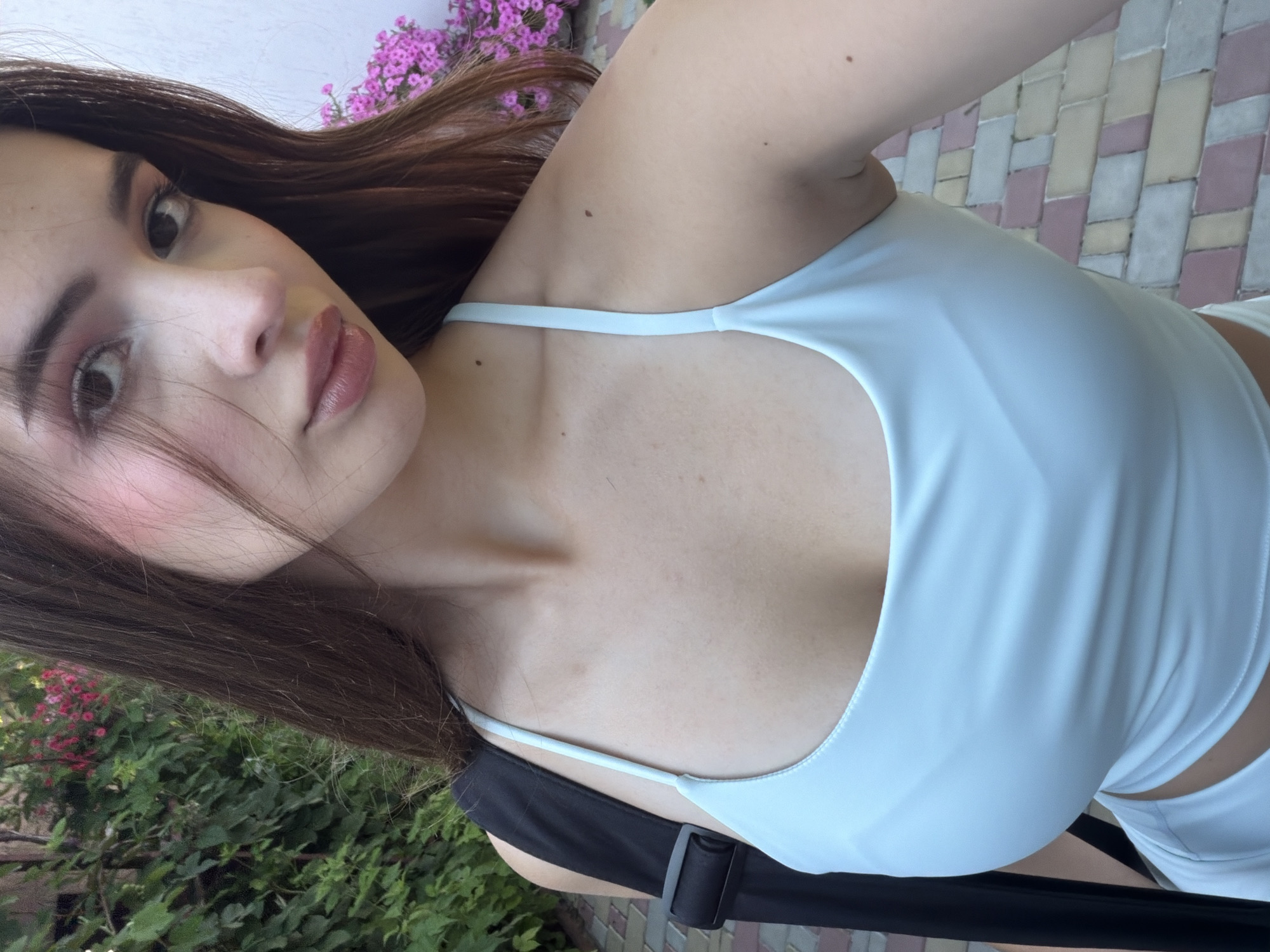 A medium-shot photo shows a young woman with long brown hair, wearing a light blue top. She is outdoors, with greenery and pink flowers in the background, and a paved walkway. The woman is looking towards the camera with her face tilted. Her makeup includes pink blush and lip gloss.