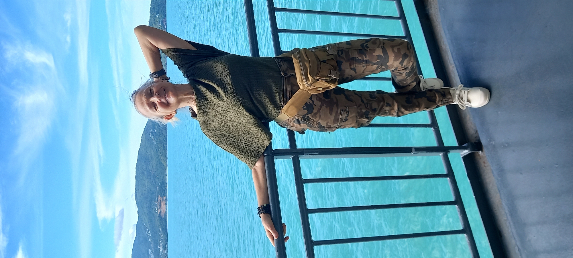 A woman stands on a metal railing structure next to a blue body of water. She has short, light-colored hair and wears a dark green, short-sleeved shirt and camouflage patterned pants. A beige bag is worn across her chest. White sneakers are on her feet. The sky is visible in the background, with land visible in the distance. This is taken in Baan Thong Po, Thailand.