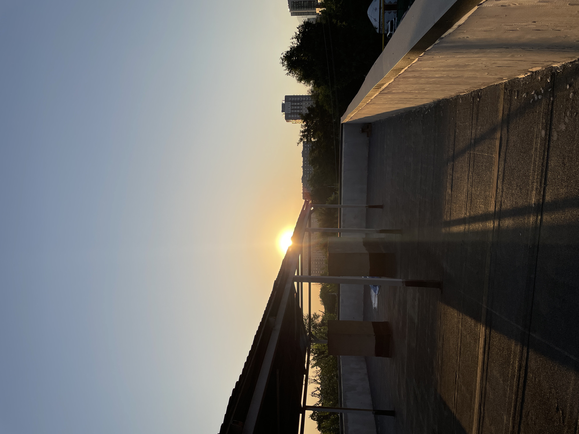 An outdoor, low-angle, medium shot captures a rooftop in Almaty, Kazakhstan, during sunset. The sun, a bright, radiant orb, peeks out from behind a covered structure on the left side of the frame, casting long shadows across the rooftop. The rooftop itself is flat and paved with dark material, showing visible lines from its construction. Two tall, rectangular structures, possibly ventilation shafts or support columns, are present on the rooftop, casting dark shadows. A white wall runs along the right edge of the rooftop, catching the warm glow of the setting sun. In the background, lush green trees and tall buildings, characteristic of an urban landscape, are silhouetted against the pale blue and yellow gradient of the sky. The overall scene depicts a calm, urban environment as day transitions into night.