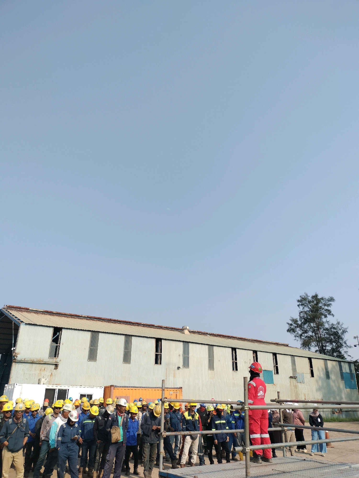 The image depicts a group of construction workers gathered outdoors in front of a long, corrugated metal building. The building appears to be industrial or warehouse-like, with several large windows and a rusted roof. In front of the building, there are shipping containers, one orange and one white. A group of individuals, most wearing yellow hard hats and work uniforms, are standing in the foreground, facing the building. One individual, dressed in a red jumpsuit and red hard hat, stands on a raised platform on the right side of the frame, appearing to address the group. The sky is clear and blue, suggesting a sunny day. The scene is likely in Batam, Indonesia, given the presence of industrial structures and a workforce common in such locations.