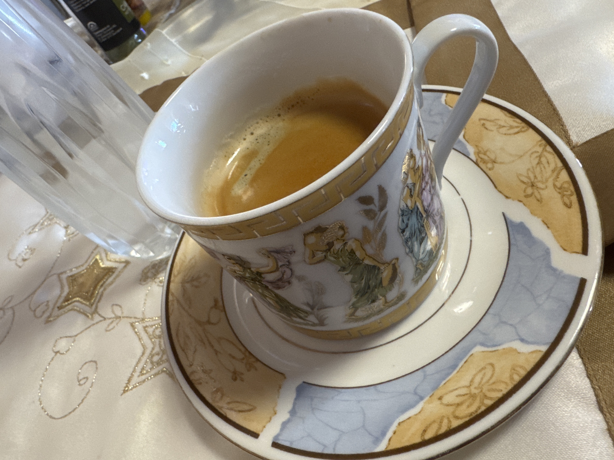 The image shows a cup of espresso on a saucer, sitting on a table with a patterned tablecloth. A clear glass bottle is partially visible in the background. The cup and saucer have a decorative design with figures and a gold trim. The tablecloth has embroidered star patterns. It is not possible to determine the location as Sofia, Bulgaria from the image alone.