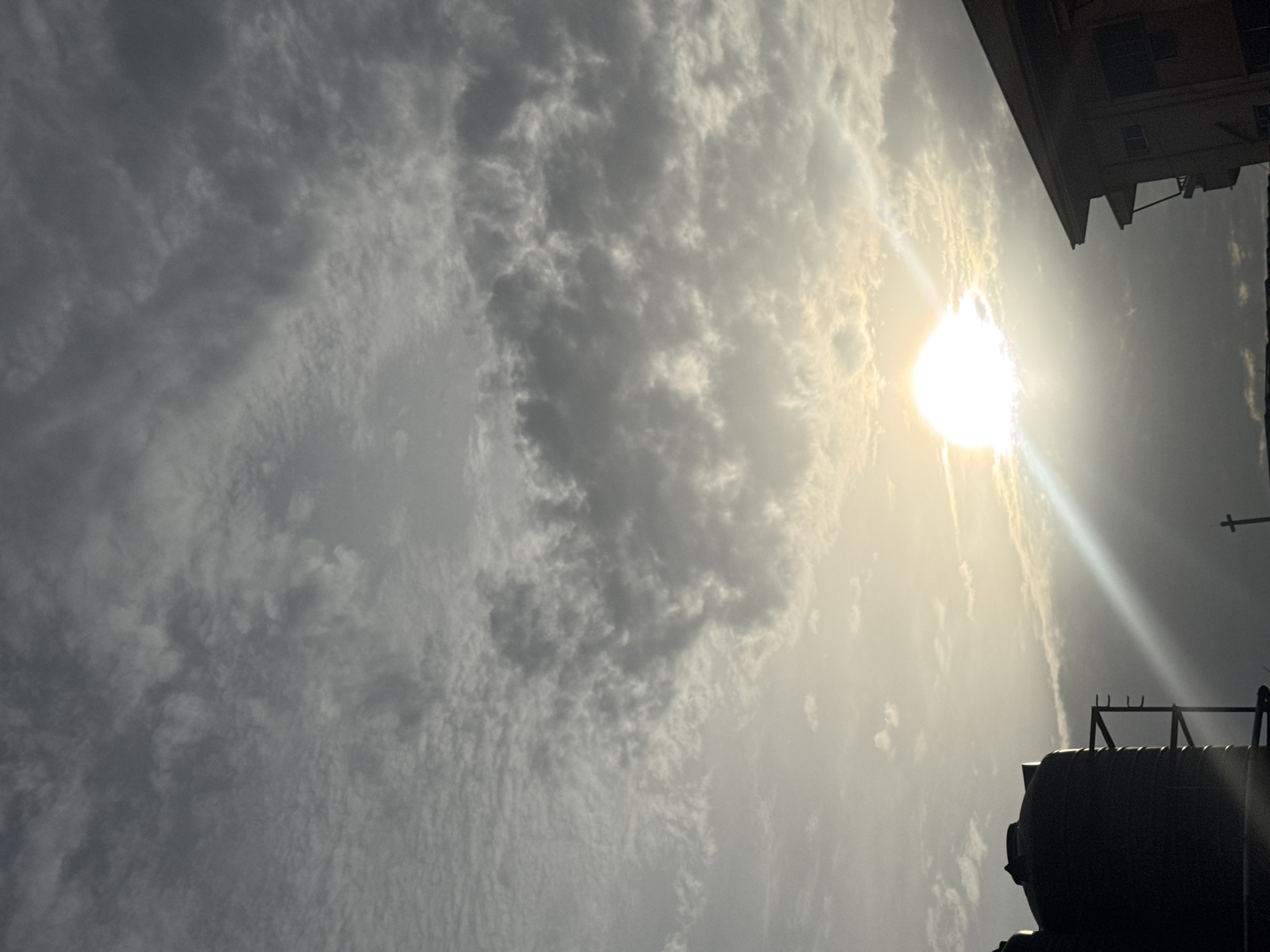 The image, rotated for viewing, depicts a sky scene in Onitsha, Nigeria. The sun is high, partially obscured by clouds, creating a bright glare with visible rays. The sky is predominantly cloudy with varying shades of gray and white. A building is visible in the upper right corner, and a large, dark cylindrical object, possibly a water tank, appears in the lower right. A power pole is to the right of the tank. The overall scene suggests a sunny day with cloud cover in an urban environment.