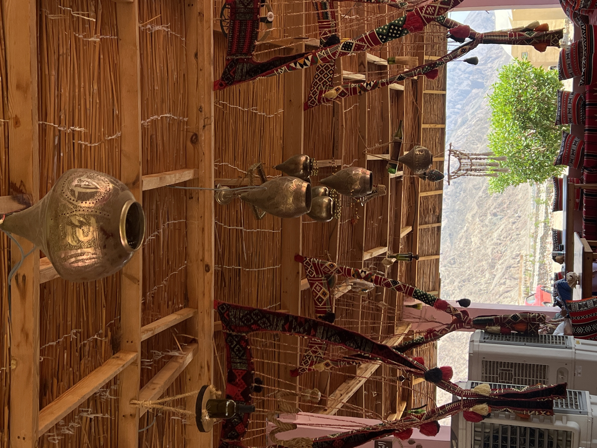 This image depicts an outdoor seating area with a rustic, traditional aesthetic, likely a cafe or restaurant, overlooking a mountainous desert landscape in an unspecified location in Egypt. The overhead structure is made of wood beams and thatched roofing. Decorative elements include several hammered metal lamps hanging from the ceiling, and colorful, woven fabric drapes with tassels and intricate patterns, possibly of Bedouin origin, are strung between poles and along the edges of the seating area. Two large, white air conditioning units are visible on the left. In the background, a lush green bush is situated in front of a rocky mountain range. A red car is parked in the distance. The seating area itself features low tables and benches adorned with cushions covered in similar patterned fabrics. A person is seated at a table on the right, facing away from the camera, with their legs crossed. The overall impression is one of a culturally rich and relaxed environment.