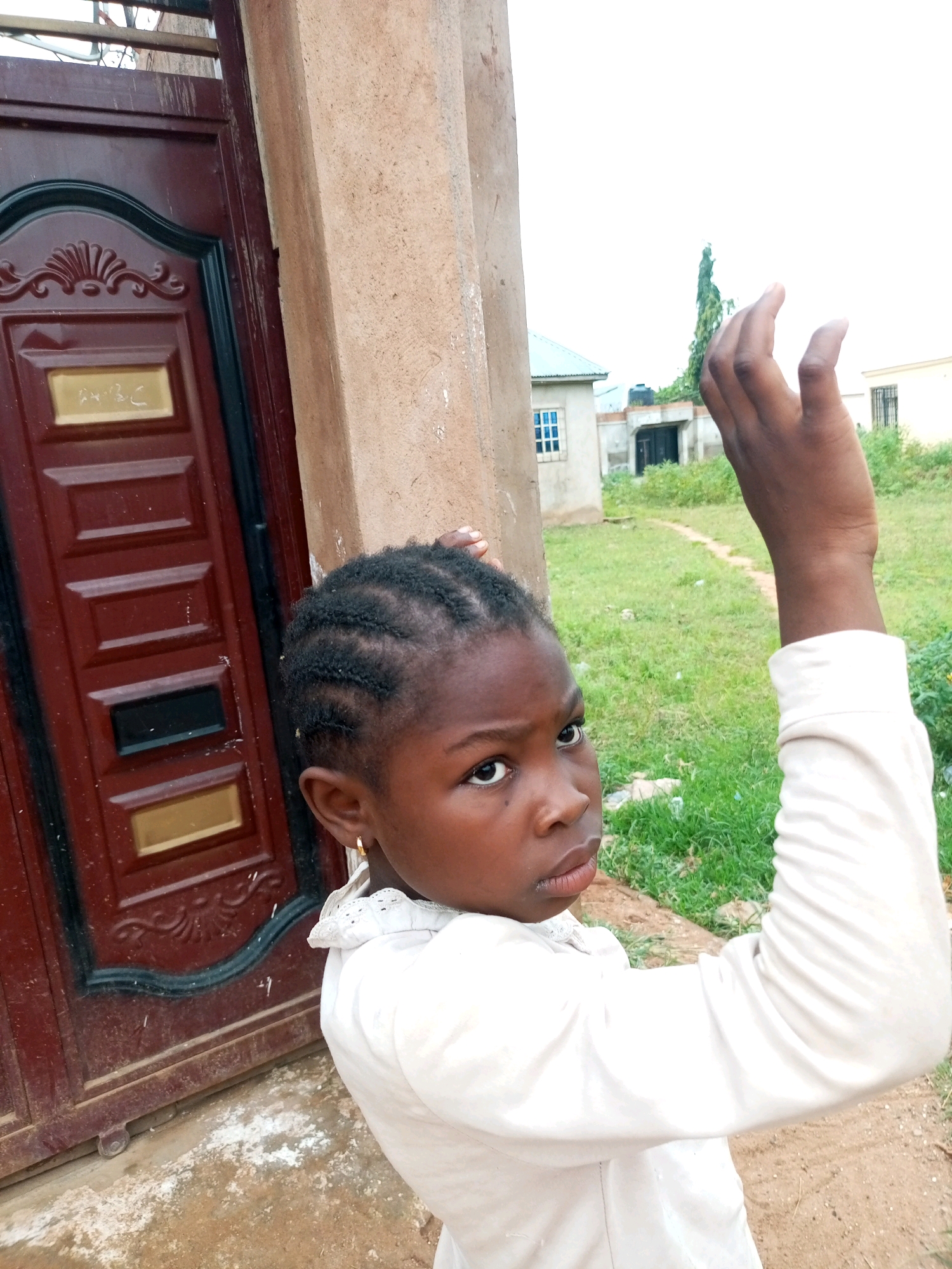 A young Black girl with braided hair stands in front of a maroon door and a tan column, her right arm raised. She wears a white collared shirt. Her left hand is on the column, and her right hand is reaching upwards, palm open. Behind her, a green grassy area is visible with some houses and a tall green tree. The sky is overcast and bright white.
