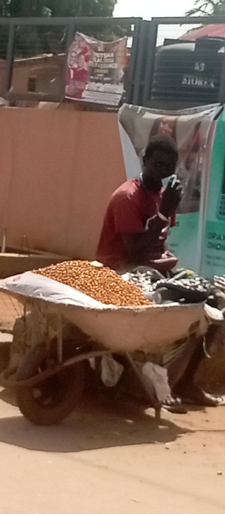 A medium shot depicts a man with dark hair sitting behind a wheelbarrow filled with peanuts in what appears to be an urban setting. The man wears a red shirt and is holding something near his mouth. The wheelbarrow, laden with a large mound of peanuts and other items, sits on a dusty surface, casting shadows. Behind the man, a partially obscured poster and what looks like a water storage tank are visible. The background shows hints of building exteriors and an overhead structure, suggesting an outdoor location in the city of Onitsha, Nigeria.