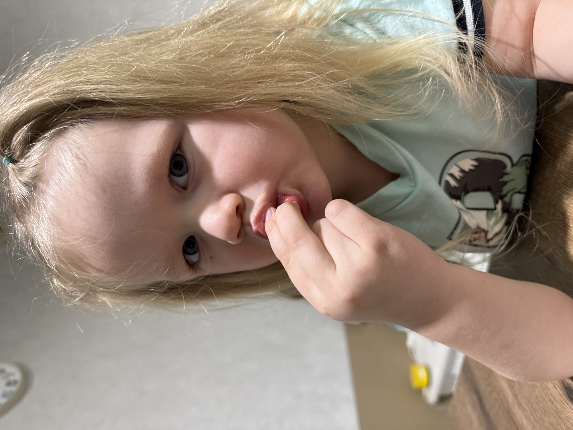 The scene depicts a young child, approximately two to four years of age, situated indoors in Gornyi (Dvurechenskoe S/P), Russia. The child, with fair skin, light blonde hair, and blue eyes, is captured in a medium close-up, looking directly at the camera. A small turquoise hair tie is visible securing a section of hair atop the child's head. The child wears a light teal long-sleeved hooded top featuring a partially visible graphic design, which includes brown and green elements, on the chest. The left sleeve displays a dark horizontal stripe with two thinner white stripes. The child's right hand is raised, with the index finger partially inserted into their mouth, contacting the lower lip and appearing to engage with something on or inside the mouth. The lips are slightly parted and moist. The child is positioned at a dark-stained wooden table with visible grain. In the blurred background, a light-colored wall is visible. To the upper left, a partially visible, blurred circular object with numerical or marking patterns suggests a clock or similar device. To the lower left background, a blurred white object with a bright yellow component on top is discernible.