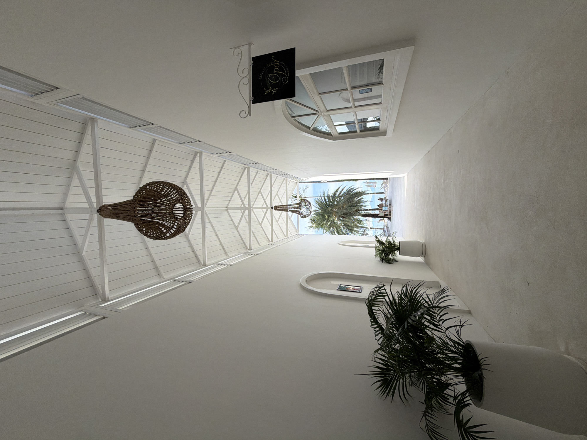 The image depicts a narrow, white-walled corridor leading towards an outdoor scene of a beach with palm trees and people. The corridor has a sloped, white-paneled ceiling with exposed white beams and two large, woven pendant lights hanging from it. The walls are smooth and white, with an arched doorway on the left and a window with a decorative arch and multiple panes on the right. A black sign with white text and a sprig of leaves hangs above the window. Large potted plants, one with dark green fronds, are placed along the left wall. The floor is a light-colored, smooth material. At the end of the corridor, a beach area is visible with lounge chairs, umbrellas, palm trees, and several people. The scene is bathed in natural light, suggesting it is daytime. The location is identified as Baan Bo Phut, Thailand.