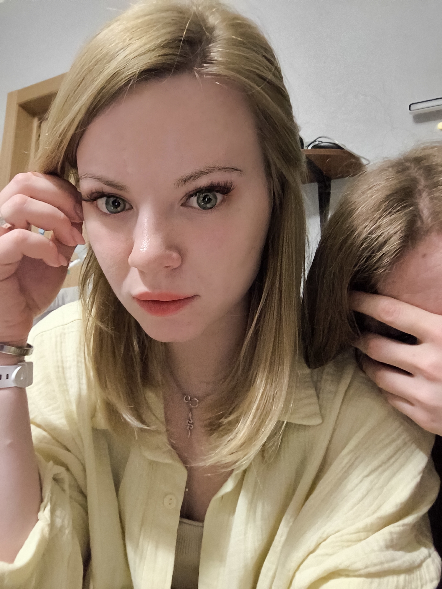 Two blonde women pose indoors. One woman is in the foreground, looking directly at the camera with one hand near her face. She wears a yellow button-down shirt and a necklace. Her skin is fair. The other woman is partially visible, partially obscured by a hand covering her face. The background includes part of a door frame and a table with a shelf above it. The location appears to be a private residence.