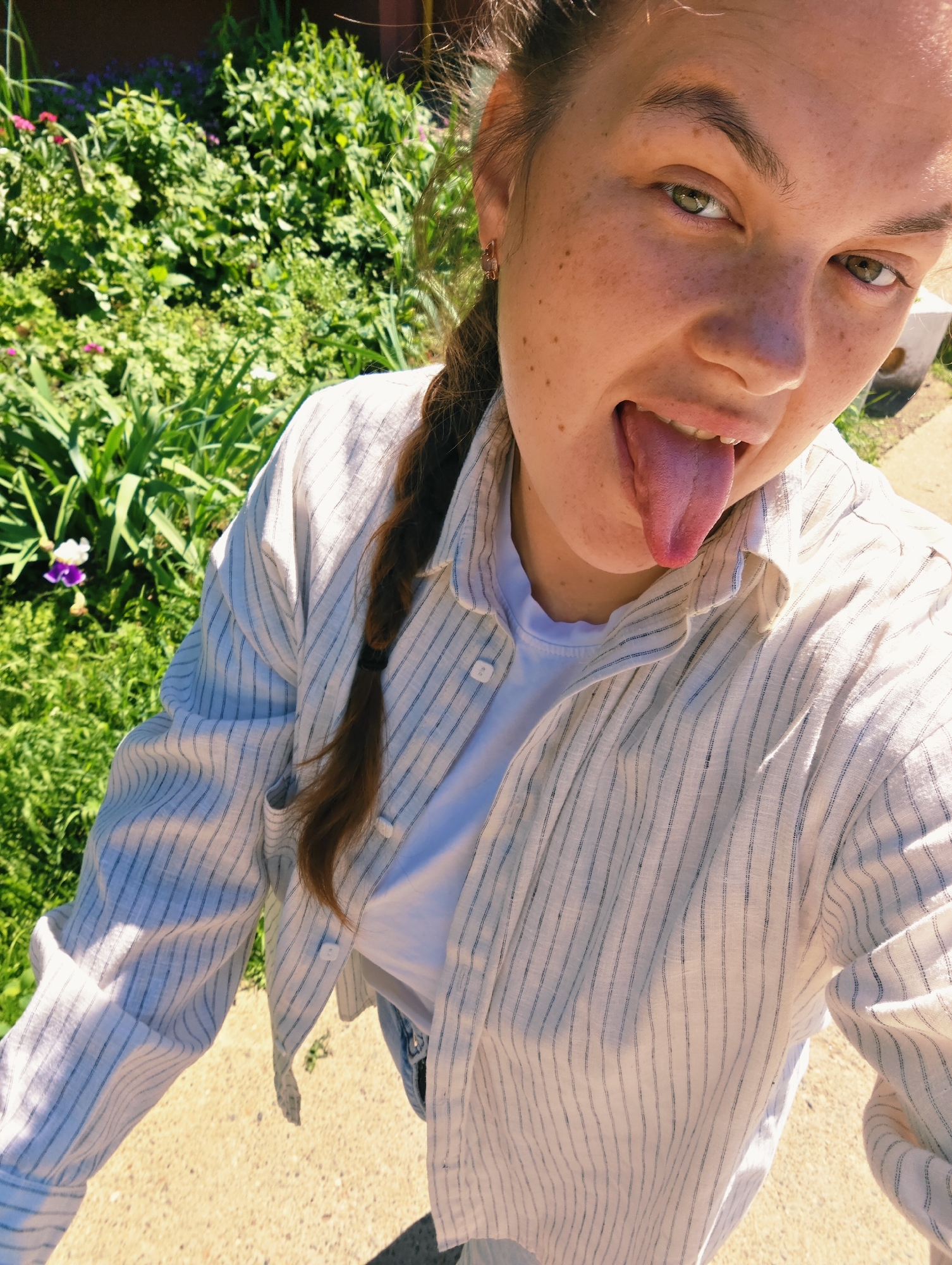 A young woman with freckles and green eyes is sticking out her tongue towards the camera, her hair in a braid. She is wearing a white t-shirt and a striped shirt over it. The background consists of lush green foliage, a pathway, and a blurry white object. The lighting suggests it is daytime. There is no indication this image was taken in Chaikovsky, Russia.