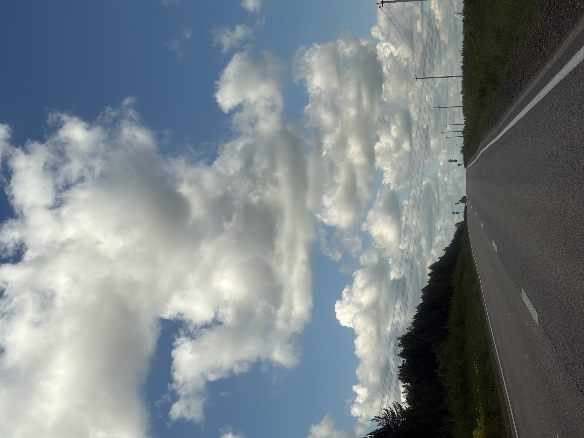 A road stretches into the distance under a partly cloudy sky. The road surface is asphalt with a white dividing line. Green vegetation lines the right side of the road. Power lines and poles are visible along the road. The location is possibly in the vicinity of Maksimovo, Russia.