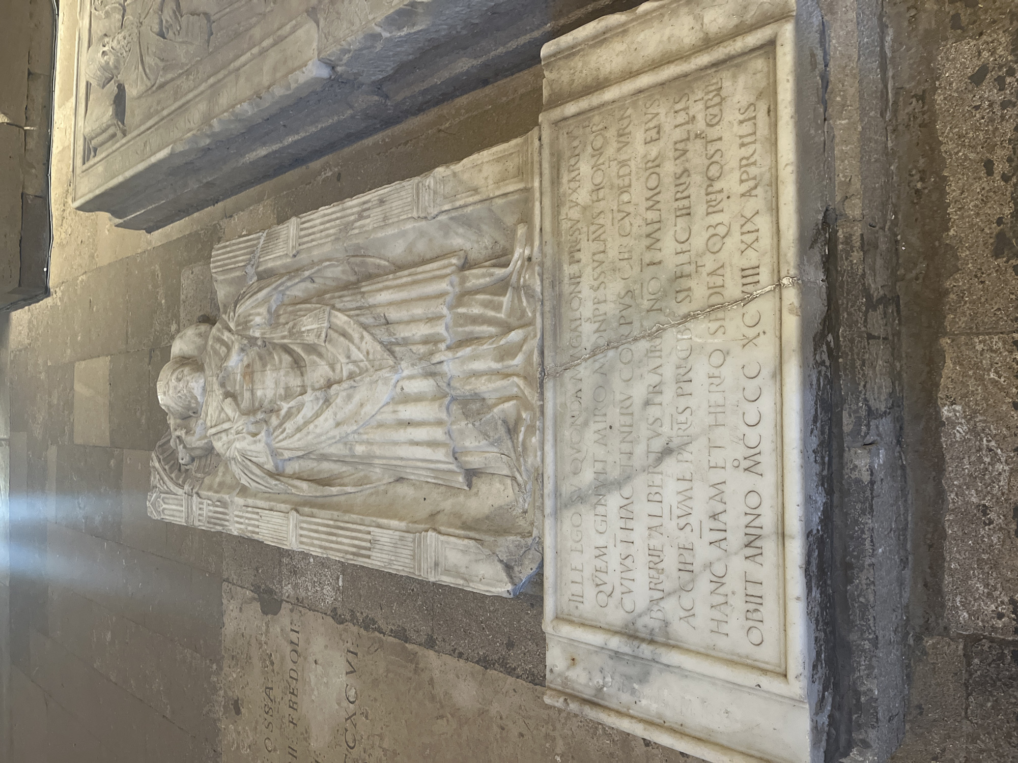 The image depicts a sarcophagus in the Cathédrale Saint-Pierre de Maguelone, located in Villeneuve-lès-Maguelone, France. The sarcophagus is made of white marble and features a recumbent effigy of a bishop or abbot, clad in episcopal vestments. The effigy is detailed, showing the individual with a mitre on his head and hands clasped over his chest. The sarcophagus rests on a stone base, and the surrounding floor is made of large stone slabs. Inscriptions are visible on the front of the sarcophagus, though the text is partially obscured by the effigy's drapery and the angle of the photograph. The lighting suggests an indoor environment, possibly illuminated by natural light shafts piercing through windows. Adjacent to this sarcophagus, another similar stone sarcophagus is partially visible, indicating a burial site within the cathedral. The overall scene conveys a sense of historical significance and solemnity.