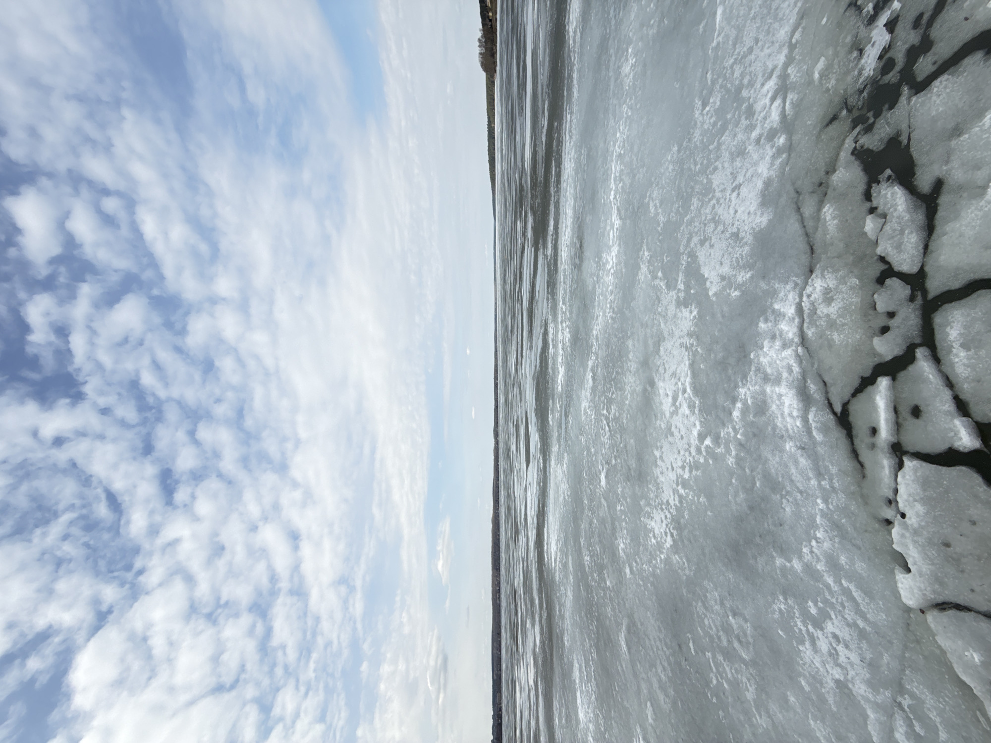 The image shows a partially frozen body of water under a cloudy sky. The right side of the image depicts ice, with cracks and patches of open dark water visible. The left side shows a blue sky with puffy white clouds. In the distance, a dark treeline is barely visible above the horizon. The location is Votkinsk, Russia. No people, actions or interactions are present.