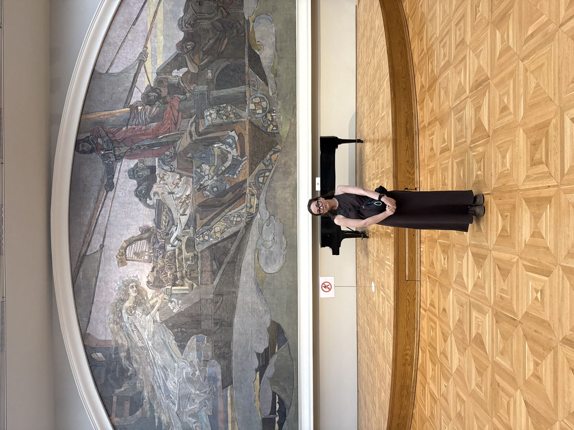 A woman stands next to a piano, in front of a large painting. The woman is wearing a dark outfit and glasses. The painting is large and ornate, depicting figures and scenes. The floor is a wooden parquet pattern. A no-smoking sign is visible. The image appears to be taken in a museum or art gallery, specifically at the Alfons Mucha Exposition in the Municipal House in Prague, not Moscow, Russia.
