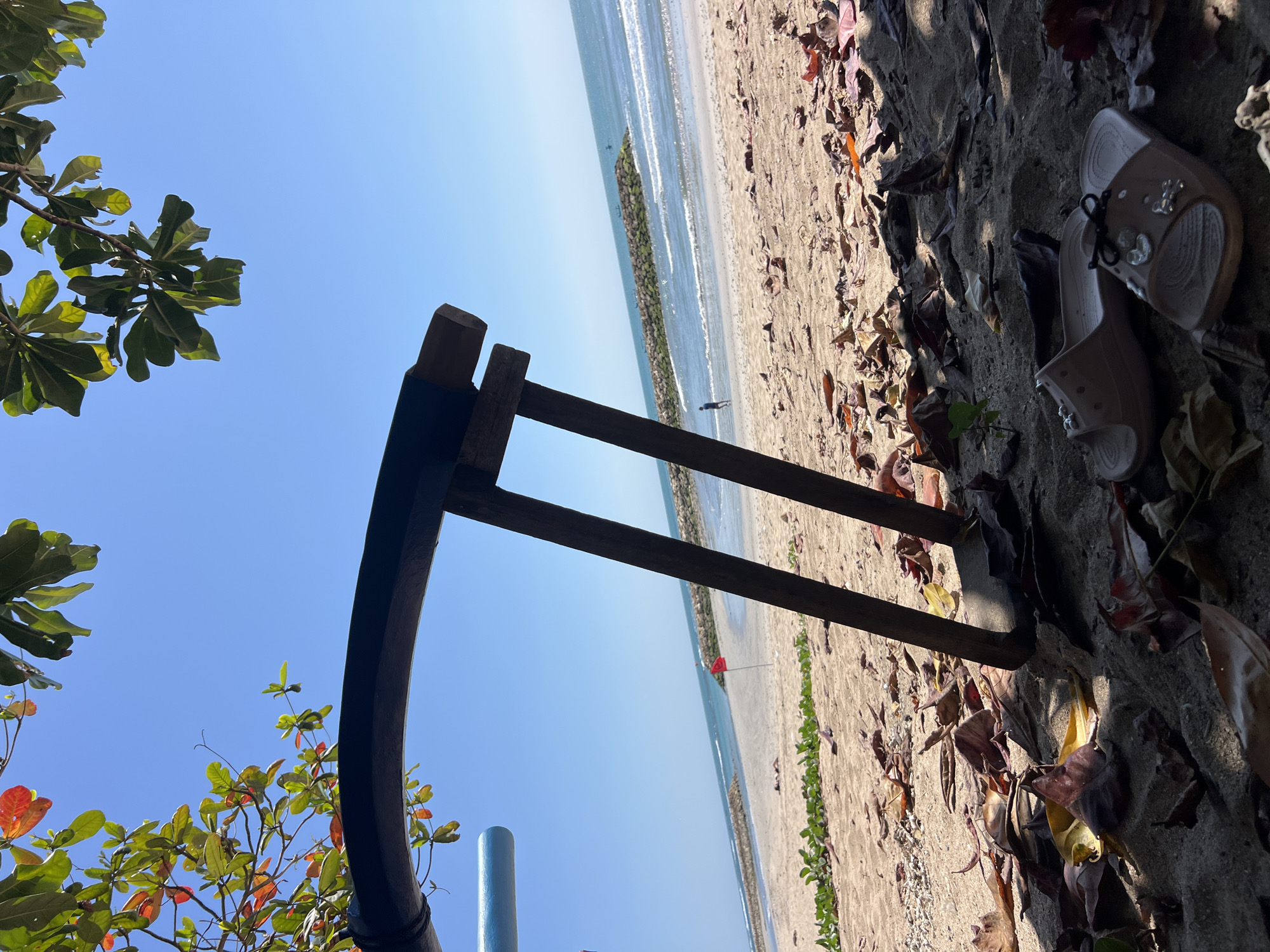 An eye-level, medium shot captures a serene beach scene in Kuta, Indonesia, with the foreground dominated by dry, fallen leaves scattered across the sandy ground. Partially visible in the lower left is a dark, curved object, possibly part of a structure or boat, and a light blue cylindrical element. Two light-colored flip-flops with decorative charms rest on the sand in the lower right. In the mid-ground, a rudimentary wooden stand with two vertical posts is positioned on the beach. Beyond the sand, the ocean stretches out under a clear blue sky, with gentle waves lapping at the shore. A stone breakwater runs parallel to the beach, with a small red flag planted on the sand near it. In the distance, a lone figure walks along the water's edge, and several small boats are visible on the horizon. Lush green foliage is visible in the upper corners of the frame, with some leaves displaying a reddish hue.