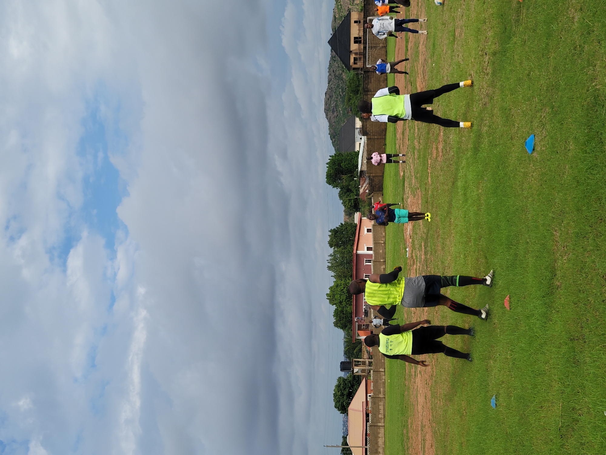 A group of men are participating in an outdoor training session on a grassy field in Jos, Nigeria. The sky is overcast with dramatic, grey clouds. In the background, several buildings and houses are visible, along with trees and rolling hills. A soccer goal is set up on the far right side of the field. Some of the men are wearing bright yellow vests and black shorts, suggesting they are part of a team or organized group. Others are in athletic attire of various colors. Some are standing, while others appear to be engaged in physical activity or drills. Small cones are scattered on the field, indicating a training exercise is in progress. The overall scene depicts a casual sports practice taking place in a semi-urban environment.