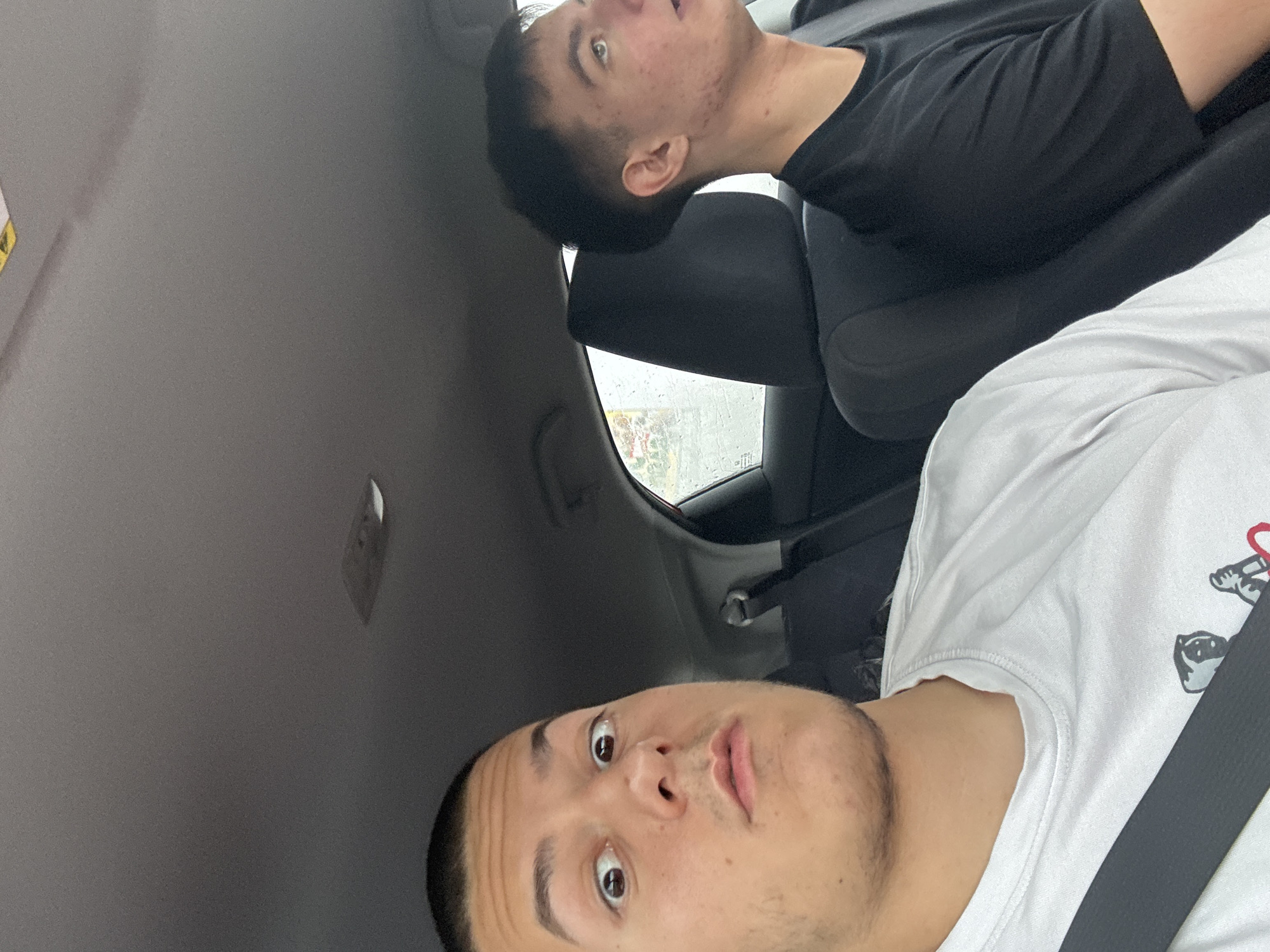 Two young men are sitting in the front seats of a car. The man in the foreground is wearing a white t-shirt and a seatbelt, looking directly at the camera with a surprised expression. The man in the background is wearing a black t-shirt and is looking upwards. The setting is indoors, likely inside a vehicle. Krustina, Bulgaria is not visibly identifiable in the image.