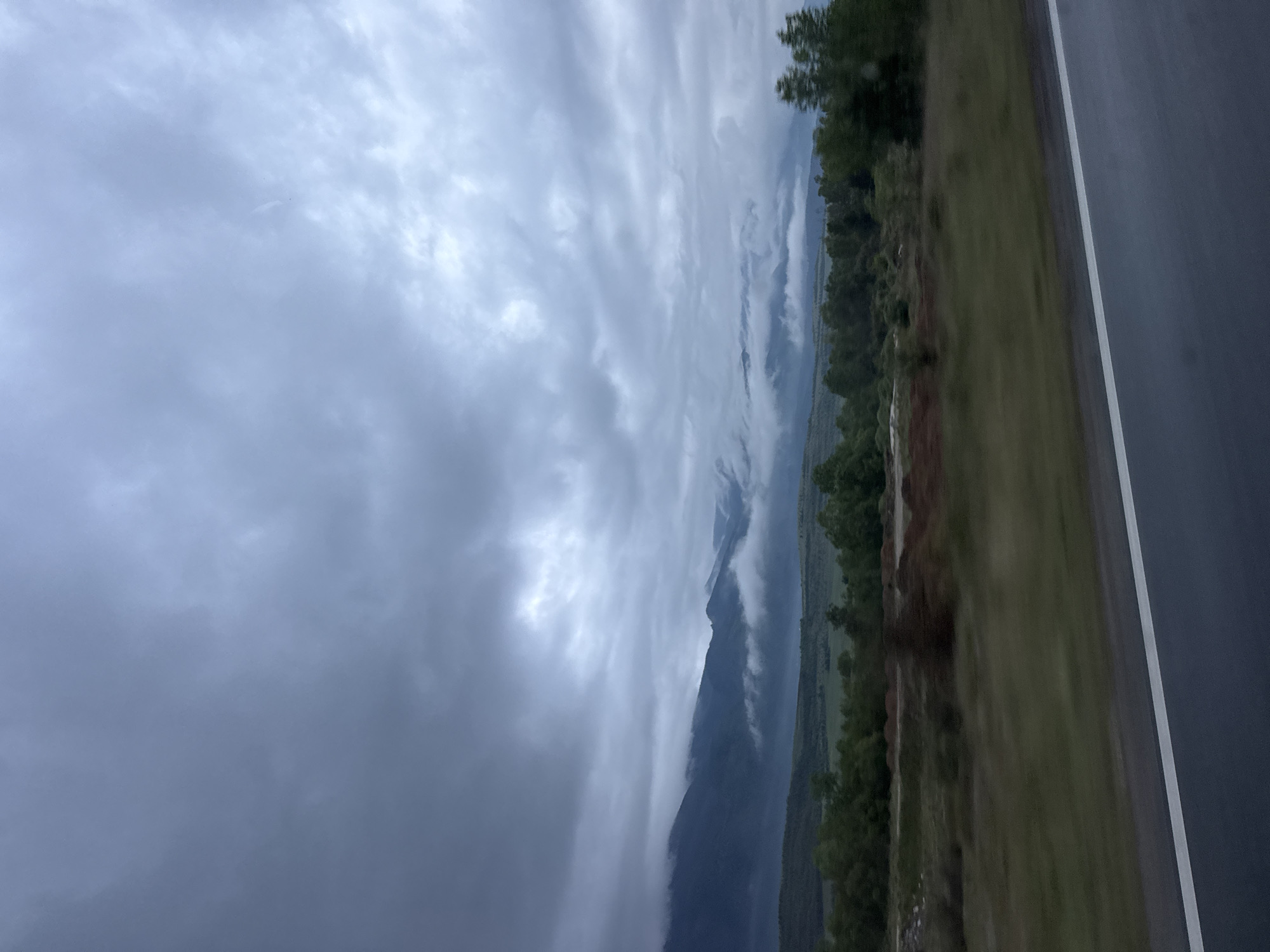 The image shows a view from what appears to be a road, with a mountain range shrouded in heavy, low-lying clouds. A paved road with a white line marking is visible on the right side of the frame. Adjacent to the road is a strip of grass and vegetation. The sky is dominated by a dense cloud cover. Based on the prompt, the location is Yahinovo, Bulgaria, implying that the mountains in the background are part of the landscape around this city.