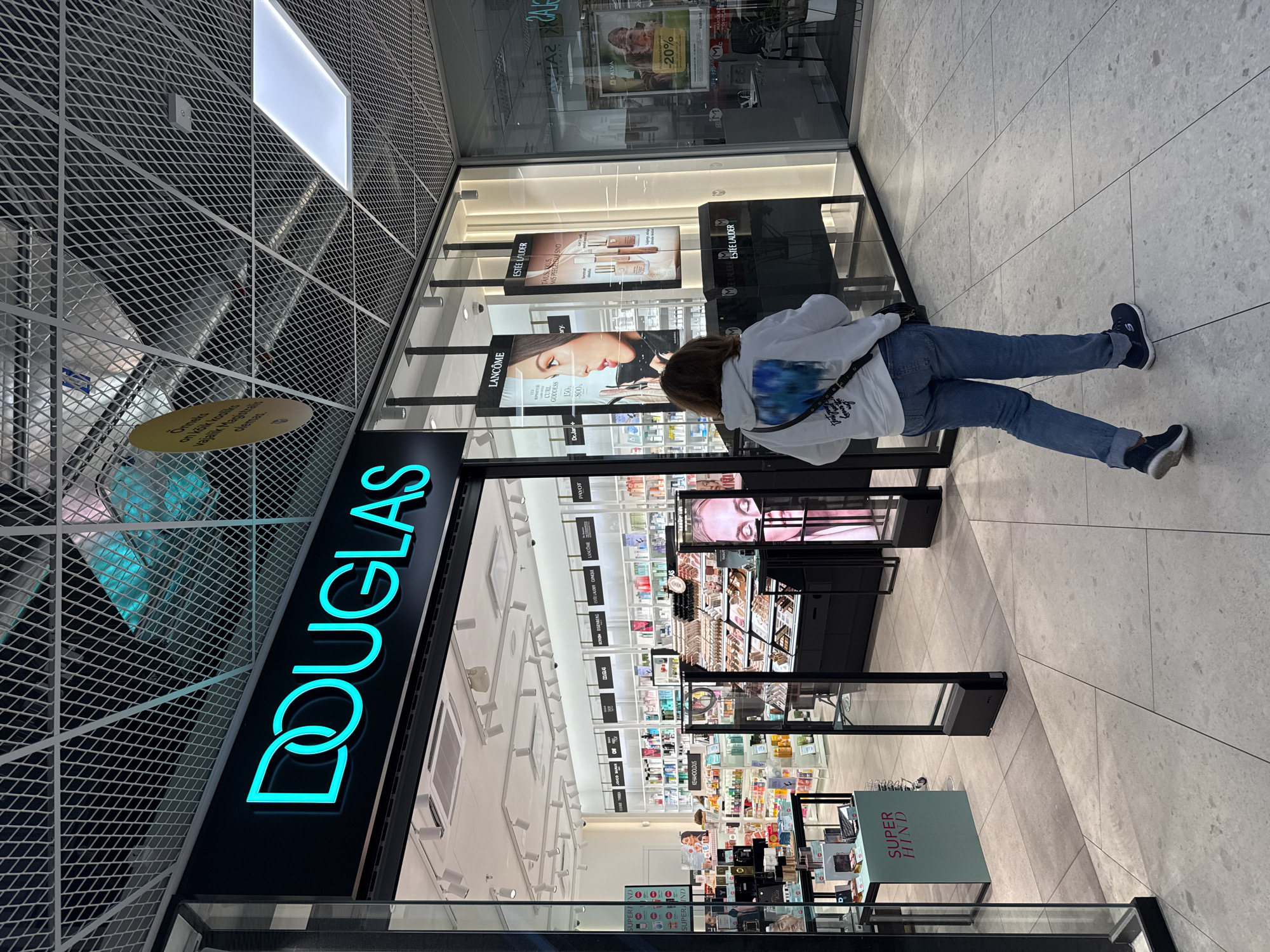 The image depicts the entrance to a Douglas perfumery store in Tallinn, Estonia. The store's prominent neon blue "DOUGLAS" sign is visible above the entrance. Through the glass storefront, rows of beauty products and displays are organized on shelves and stands. A woman wearing a light-colored hoodie and blue jeans is walking away from the camera, entering the store. The floor is tiled in a light gray stone pattern, and a metal grid ceiling with lighting fixtures is overhead. Promotional displays for brands like Lancôme and Estée Lauder are visible on the right side of the entrance. The overall environment suggests an indoor shopping mall setting.