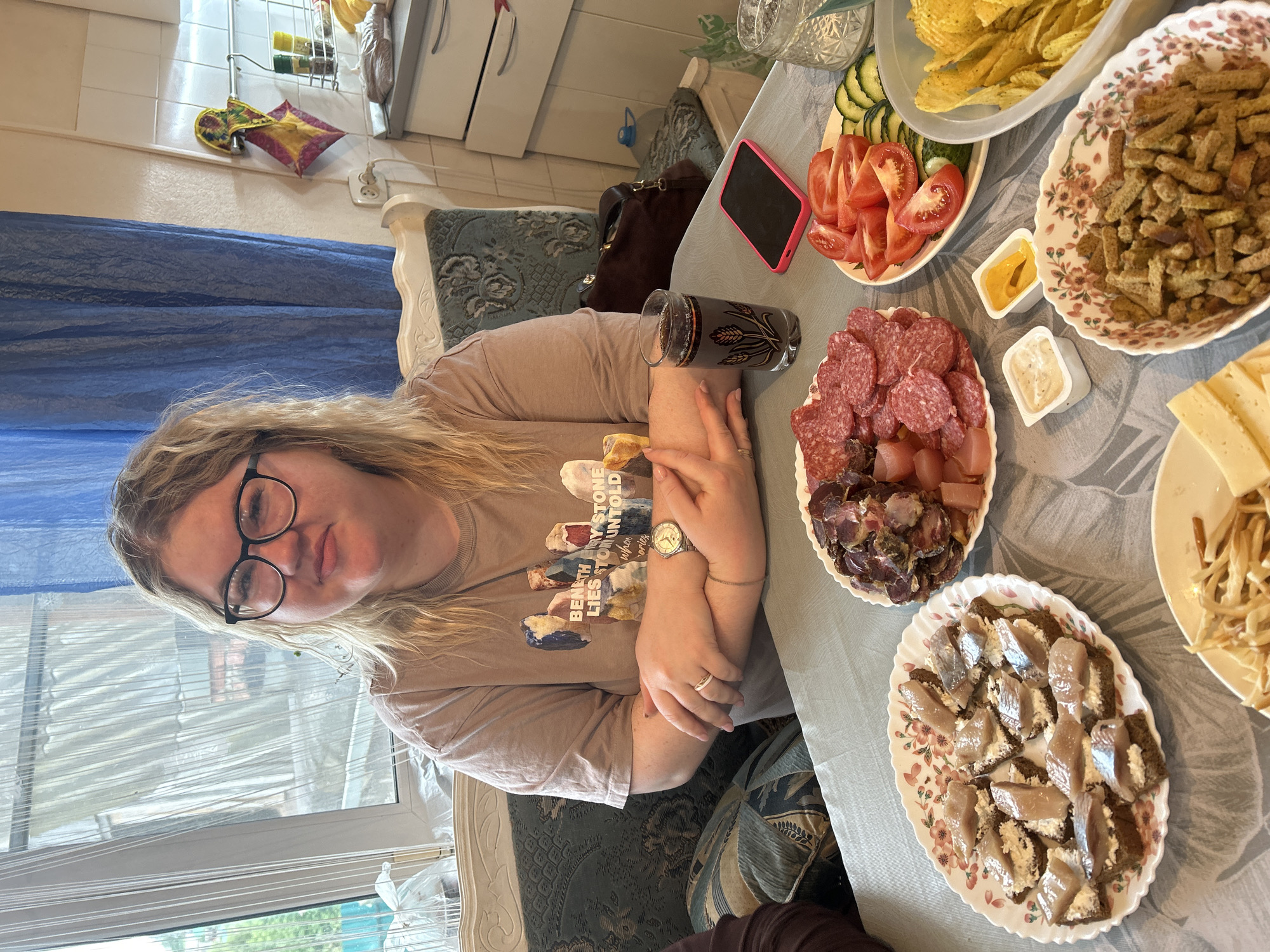 A woman with blonde hair and glasses is seated at a table laden with food. She is wearing a beige t-shirt and a watch. The table holds various plates of food including chips, crackers, sliced tomatoes and cucumbers, salami, cured meats, cheese, and open-faced sandwiches with fish. There's also a glass of dark liquid, a smartphone, and small containers of sauce. The setting appears to be indoors, possibly a home. No specific information confirms the city Фоки, Russia.