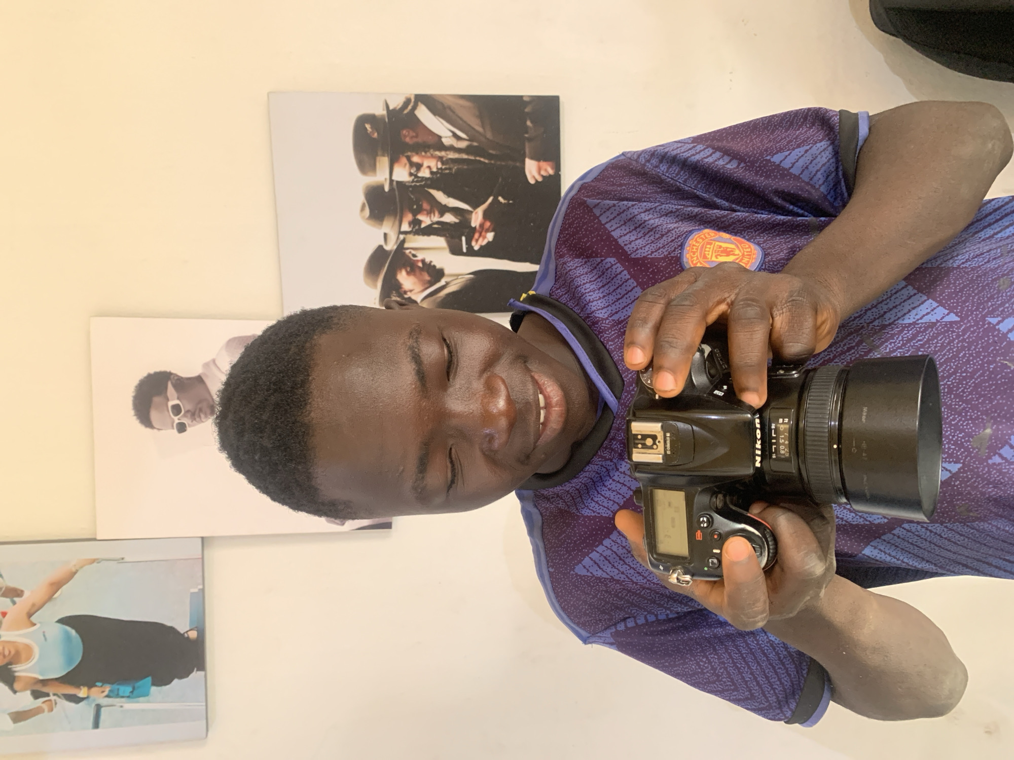 A young man with dark skin and short dark hair is captured smiling broadly while holding a black Nikon DSLR camera. He is wearing a short-sleeved purple t-shirt featuring a subtle blue patterned design and a prominent red and gold Manchester United Football Club crest on his right chest. His hands are positioned on the camera body and lens, suggesting he is either operating the device or posing with it.
The camera clearly displays the "Nikon" brand name. The attached lens, a "AF-S Micro Nikkor 60mm 1:2.8G ED," has visible focus distance markings such as "0.8 1," "1.5 5," "3," "10," and "∞." Both the camera's top and rear LCD screens are visible, showing various settings and numbers, though the specific details are not clearly legible.
The background is a light-colored wall adorned with three prints or posters. The top right print features three men in suits and fedora-style hats, with prominent dreadlocks, appearing in an artistic or promotional shot. To the left and slightly below this, another print shows a person with dark skin wearing white sunglasses, captured in a profile. The bottom left print depicts a woman in a light blue top and dark pants, posed in an active or dance-like manner.
The man's joyful expression indicates enthusiasm or contentment. This image is set in Jos, Nigeria.