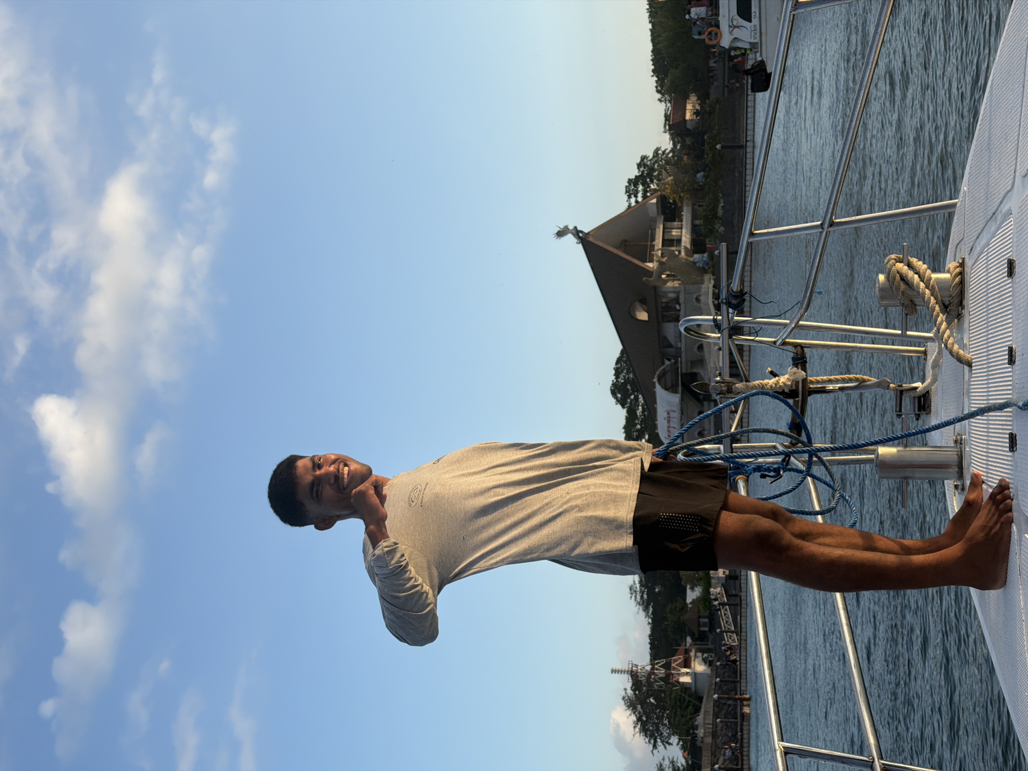 A full shot shows a young man with dark skin and short black hair standing on the deck of a white boat, looking to his right with a wide smile and his right hand raised in a peace sign. He is wearing a long-sleeved grey t-shirt and black shorts. The boat is on the water, with a railing in the foreground. In the background, there are buildings with pointed roofs, lush green trees, and a blue sky with scattered white clouds. The scene appears to be a sunny day, possibly in the late afternoon given the warm light. The man is the primary subject, and his relaxed posture and cheerful expression suggest a leisurely activity, likely a boat trip. The background hints at a coastal or waterfront area within a city, possibly Denpasar, Indonesia, given the architectural style of the buildings and the tropical vegetation.