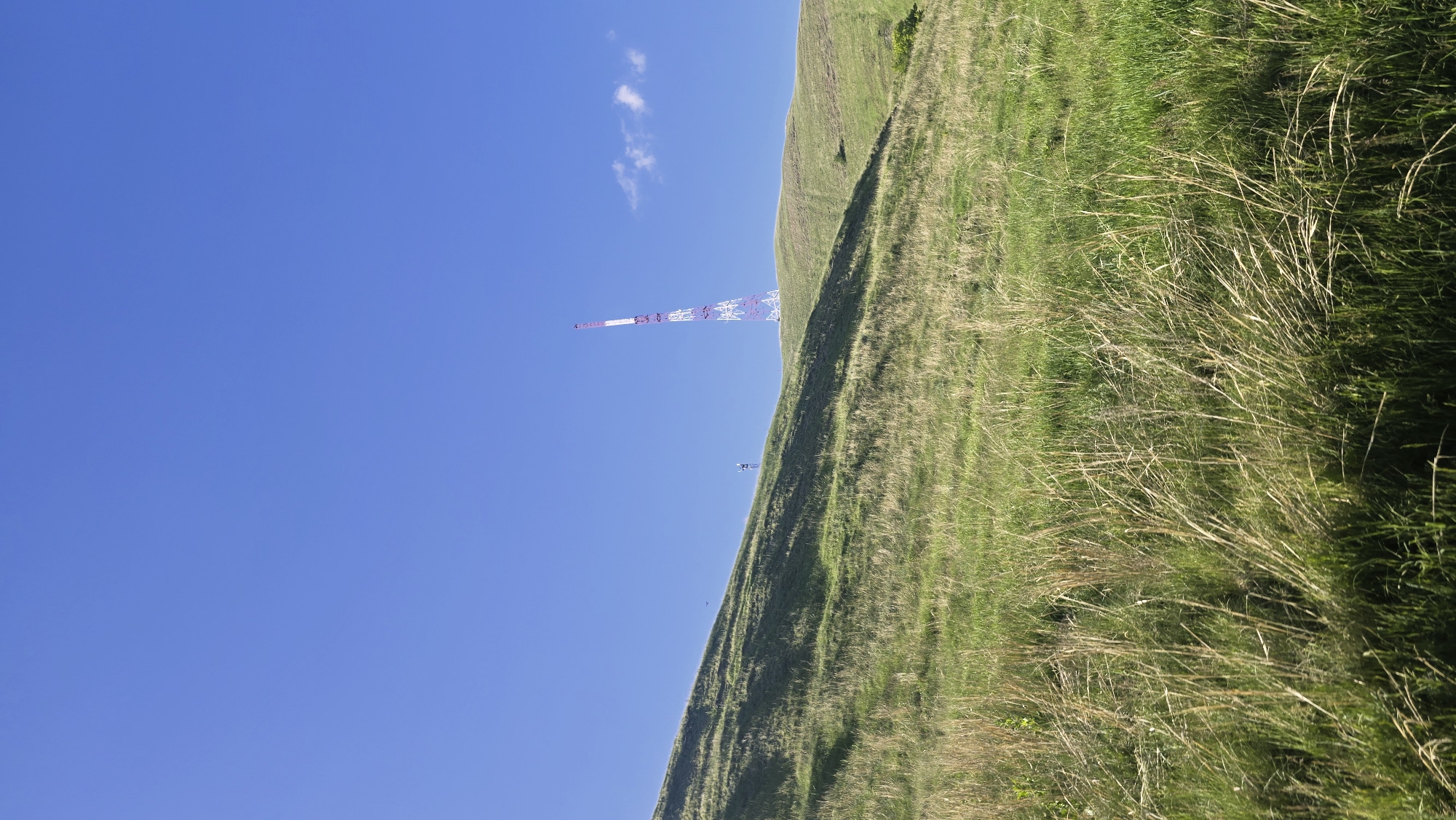 A tall, slender radio tower with red and white sections stands prominently on a grassy hilltop under a clear blue sky. The tower is located in the city of Karabash, Russia. The foreground of the image is filled with tall, dry grass, creating a textured contrast with the smoother, green slopes of the hill behind it. A small, distant figure can be seen standing on the hillside, possibly observing the tower or the landscape. Another, smaller tower is visible further in the distance on the left side of the frame. A few wispy clouds are scattered in the sky, adding a subtle detail to the otherwise expansive blue. The overall scene depicts a vast, natural landscape with a man-made structure dominating the horizon.